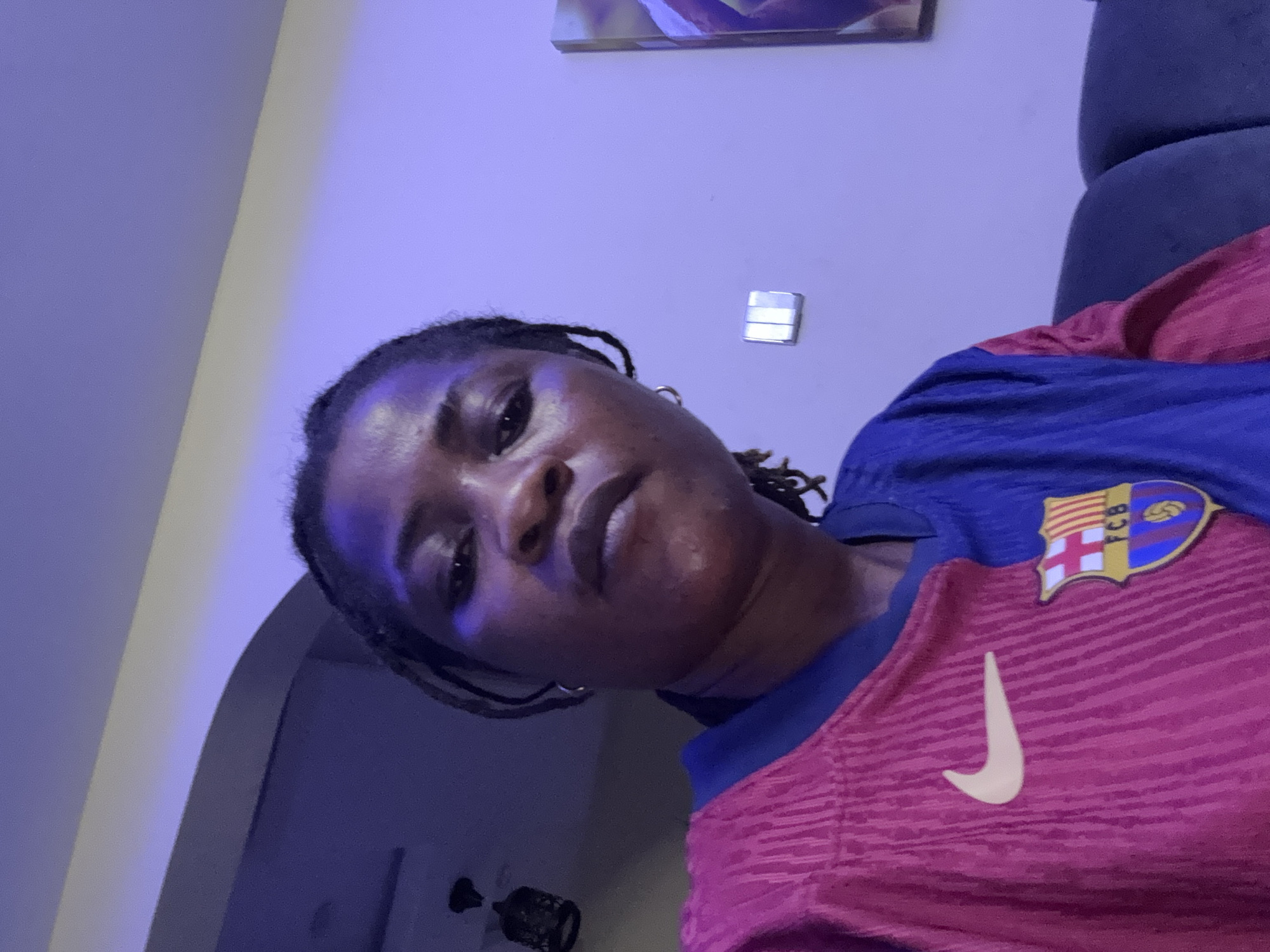 The image is a close-up, selfie-style portrait of a woman with dark skin and braided hair, wearing a pink and blue soccer jersey. The jersey features a yellow Nike swoosh on the left side and the FC Barcelona crest on the right side. The background appears to be an indoor setting with a light purple hue, possibly due to ambient lighting or a colored wall. A light fixture is visible in the upper left corner, and a framed picture hangs on the wall to the right. The woman is looking directly at the camera with a neutral expression. There are no visible indications of the specific location within Ajara, Nigeria, other than it is an indoor domestic setting.