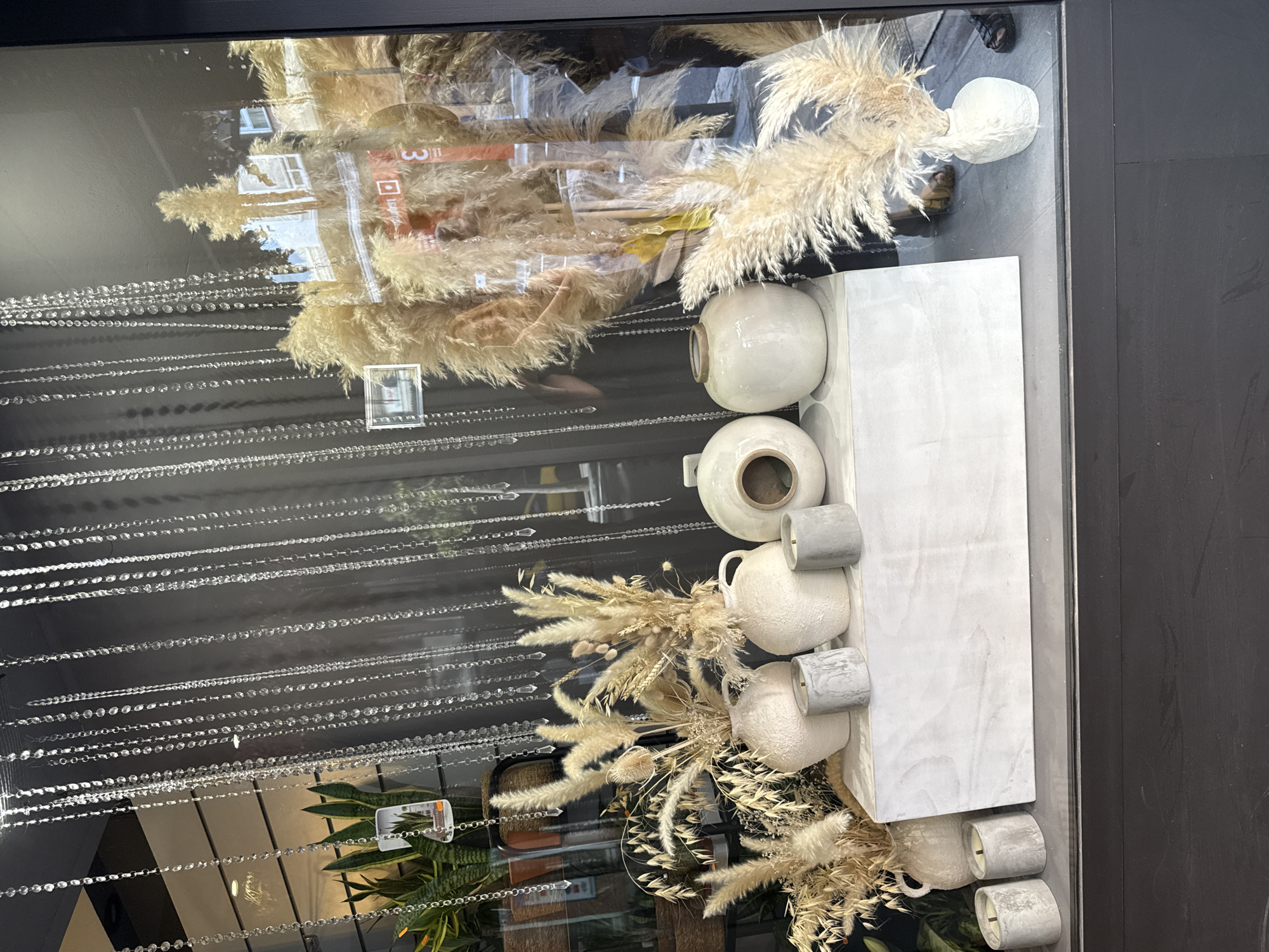This media file captures a shop window or interior display, featuring a blend of natural and minimalist elements. The image is taken through glass, which creates prominent reflections of the surrounding environment.
The main subjects of the display are dried floral arrangements, predominantly composed of fluffy pampas grass and various other dried stalks in earthy tones of beige, cream, and light brown. These arrangements are artfully placed within and around several textured, off-white or cream-colored ceramic vases and jugs of different shapes and sizes. Alongside the pottery, several small, cylindrical objects, likely candles, with a grey, concrete-like finish are arranged on a light-colored, possibly grey or white, rectangular display platform.
In the foreground of the display, hanging vertically, are multiple strands of decorative chains made of clear, reflective beads or crystals, adding a touch of sparkle and texture.
The reflections on the glass are a significant part of the image, showing glimpses of the exterior or opposite interior. These reflections include parts of a building with windows, suggesting an urban setting or a well-lit indoor space. Greenery, specifically what appears to be snake plants in pots, is also visible in the reflections. Faint human presence is suggested by a reflection of feet wearing sandals. A blurred, mirrored numerical digit "3" or "w" and a square icon are also discernible in the reflections, possibly from a sign or advertisement outside.
The overall aesthetic of the display is modern, natural, and calm, indicative of a home decor store, florist, or boutique. There is no discernible activity taking place, as it's a static display. The lighting suggests daytime or a brightly lit interior.