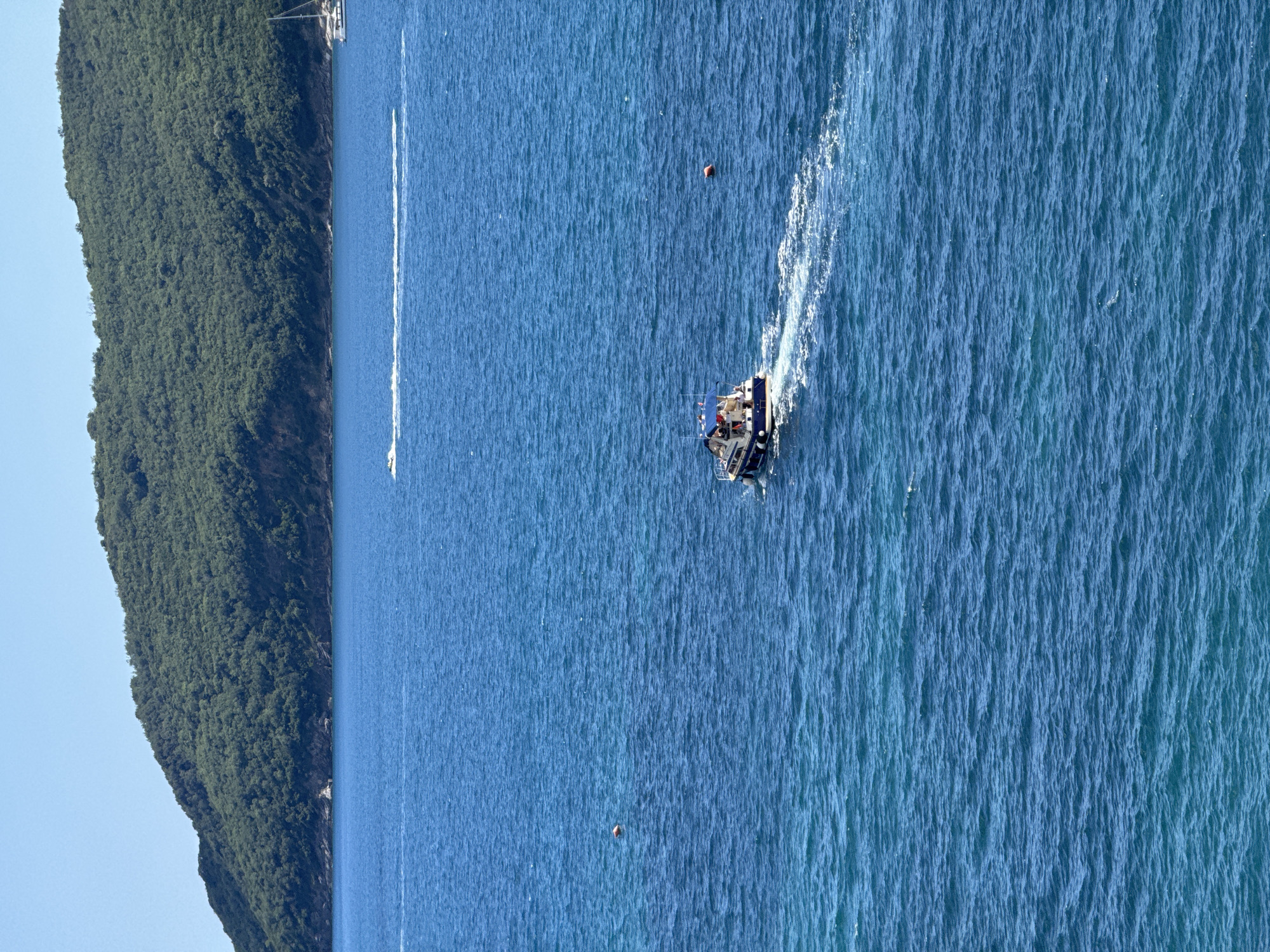 A medium shot captures a clear, bright day with a vast expanse of blue water. In the foreground, a white and blue motorboat with several people on board travels away from the viewer, leaving a white wake behind it. The boat has a canopy over a portion of its deck. In the middle ground, a red buoy floats on the water. Further in the distance, a jet ski creates a long white spray trail across the blue surface. The background is dominated by a large, lush green hillside that slopes down to the water's edge. The hillside is densely covered with trees and vegetation. The sky above is a clear, pale blue. The scene appears to be a view of the Adriatic Sea, likely near Budva, Montenegro, given the characteristic mountainous coastline.