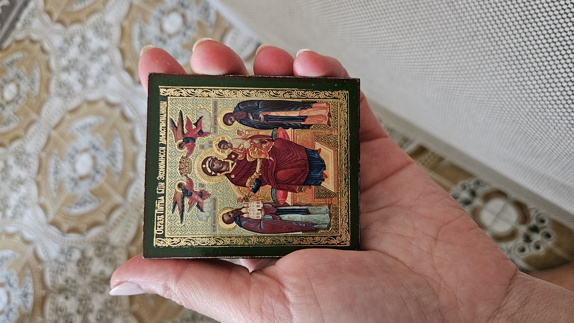 An antique religious icon is held in a person's hand. The icon is square and depicts the Virgin Mary holding the Christ Child, flanked by two saints and two angels. The background of the icon is gold. The icon is framed by a dark green border. The person's hand is holding the icon with their thumb and index finger on the left side, and their middle, ring, and pinky fingers on the right side. The background of the image is a patterned textile. There is no discernible location in the city of Mondolfo, Italy.