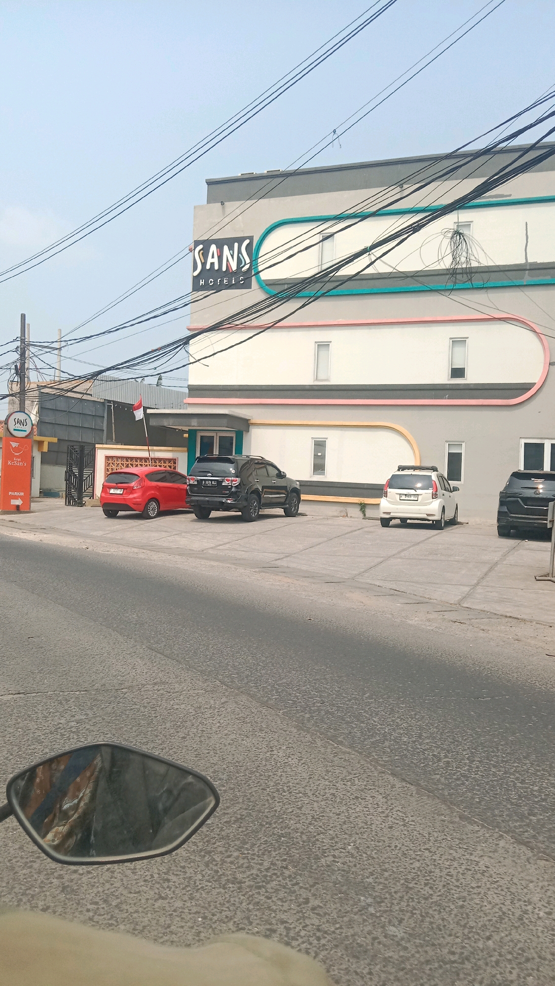 The image shows the exterior of a SANS Hotel in Bekasi, Indonesia. The hotel is a multi-story building with a modern design, featuring horizontal bands of color in teal and pink. Several cars are parked in front of the hotel, including a red hatchback, a black SUV, a white hatchback, and a dark-colored SUV. A sign for "Kopi KeSan's" and "PARKIR" is visible near the entrance. The sky is clear and blue. Overhead power lines are visible across the top of the image. A red and white Indonesian flag is flying on a pole. The scene appears to be a sunny day.
