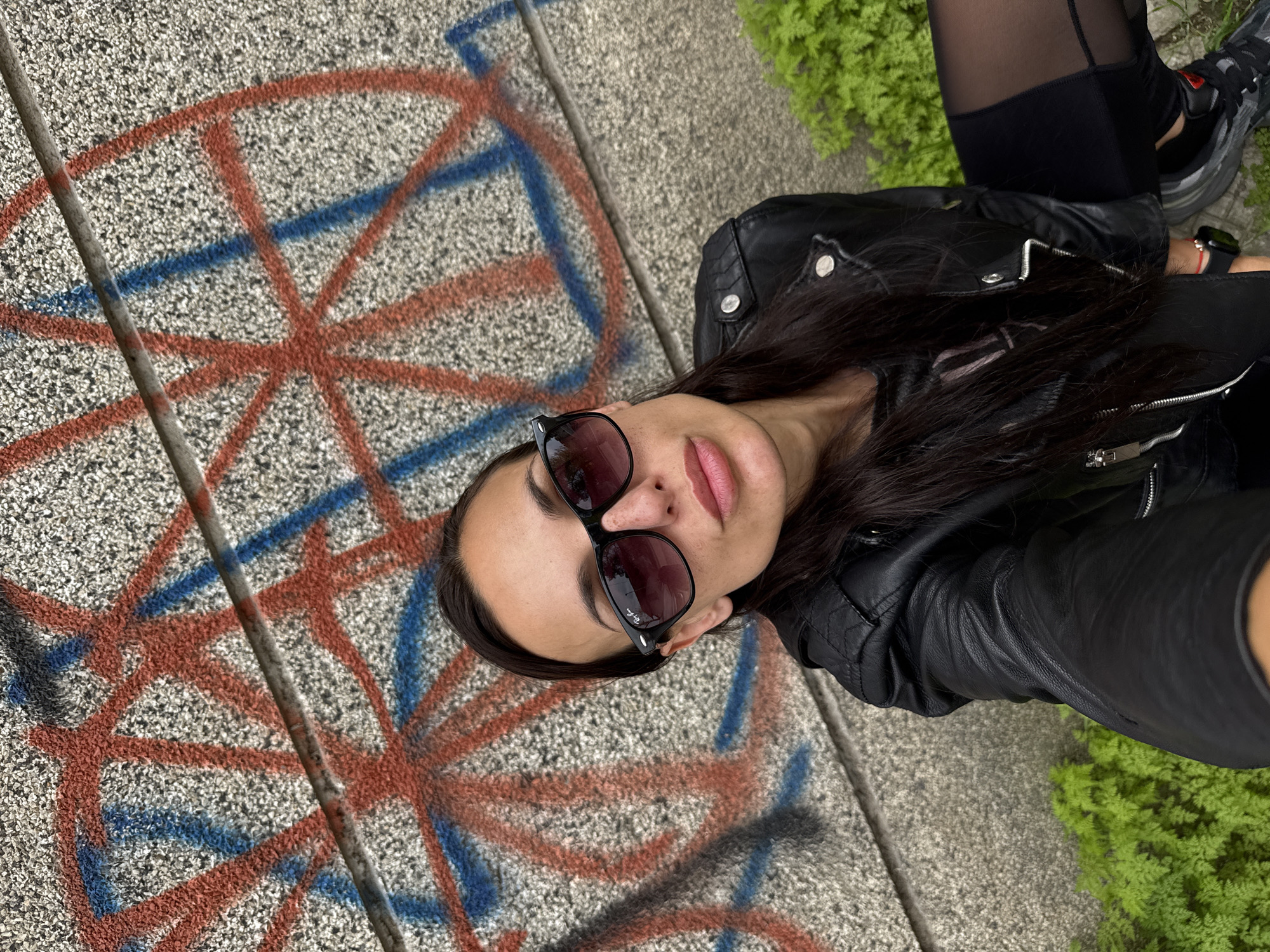 A woman is posing for a selfie next to a concrete surface marked with red and blue graffiti. She is wearing sunglasses and a black leather jacket. Part of her lower body is visible, she is wearing black leggings and black and grey sneakers. Green foliage is seen next to the concrete surface. The location is in the city of Pleven, Bulgaria.