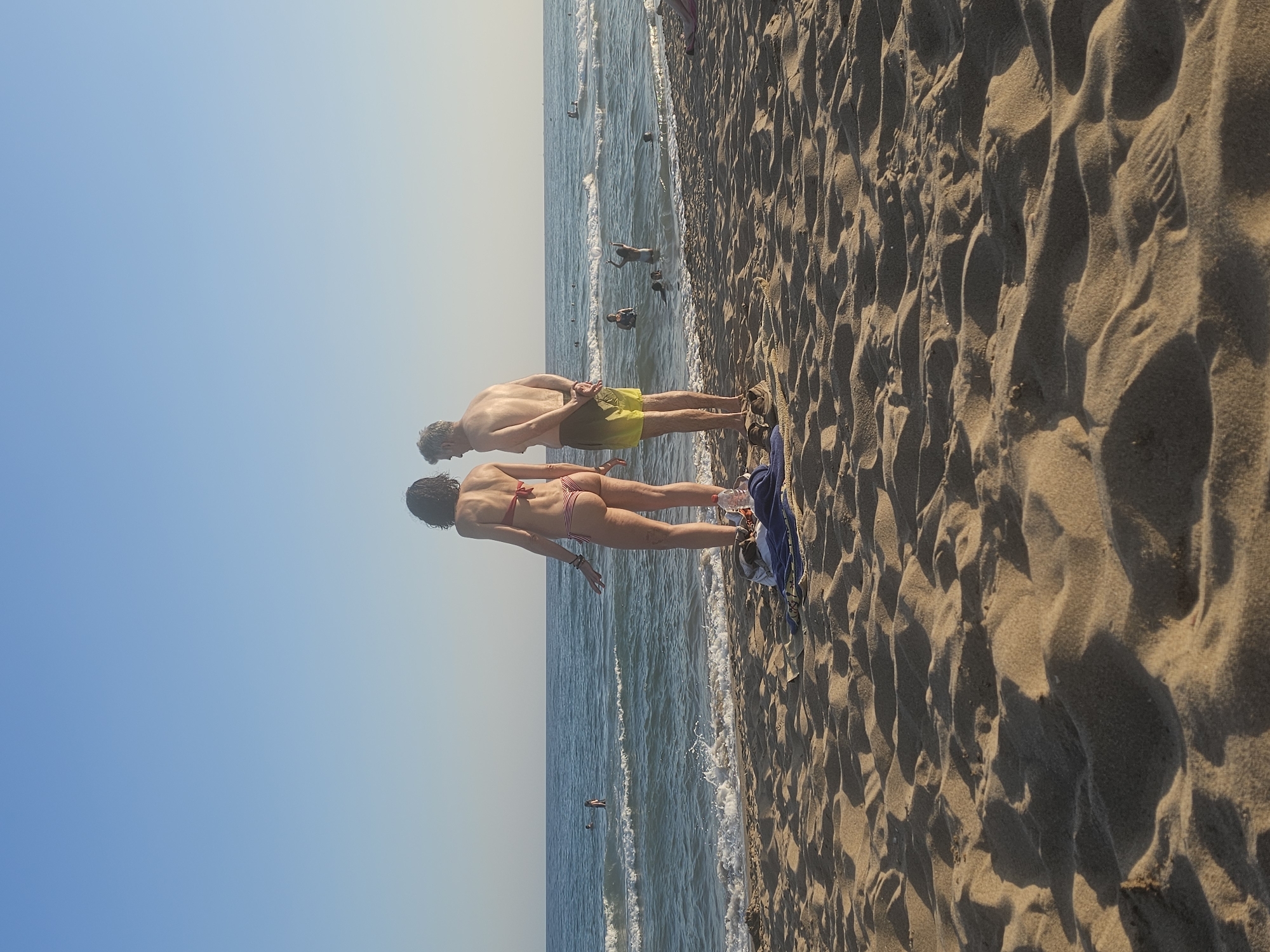 Two individuals, a man and a woman, are standing on a sandy beach facing the ocean. The man is wearing yellow shorts, and the woman is wearing a red and white striped bikini. They are positioned close to the water's edge, with the man slightly behind and to the left of the woman. In the background, several other people are visible in the water, and some birds are scattered on the sand and in the sea. The beach is covered in textured sand with visible footprints and ripples. The sky is a clear, pale blue, transitioning to a lighter hue near the horizon, suggesting it is daytime. The scene depicts a relaxed beach atmosphere, likely a warm day, given the attire of the individuals. The orientation of the image suggests it was taken in portrait mode and possibly rotated. No visible text is present in the image. The location context is Castelldefels, Spain.