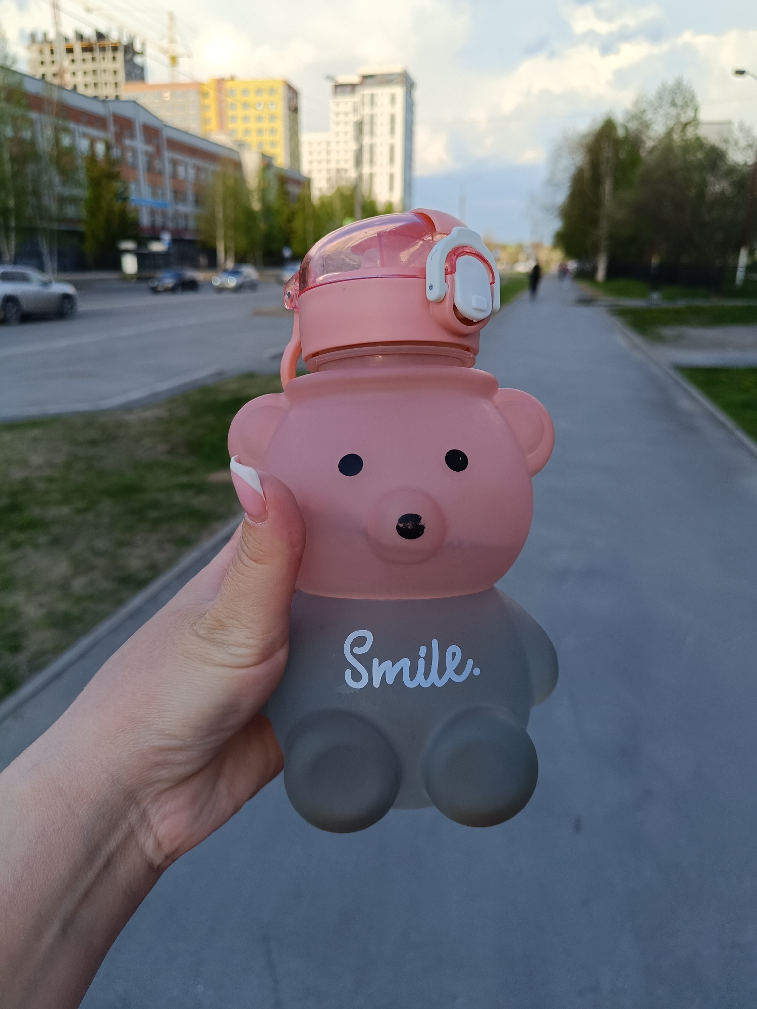 A person holds a pink and gray plastic water bottle in the shape of a bear. The bottle has a pink lid and the word "Smile" written on its gray body. The person's hand and arm are visible. The background shows an urban scene with a wide sidewalk, a street with cars, buildings, and trees. The location is likely Tobolsk, Russia based on the architecture and urban layout.