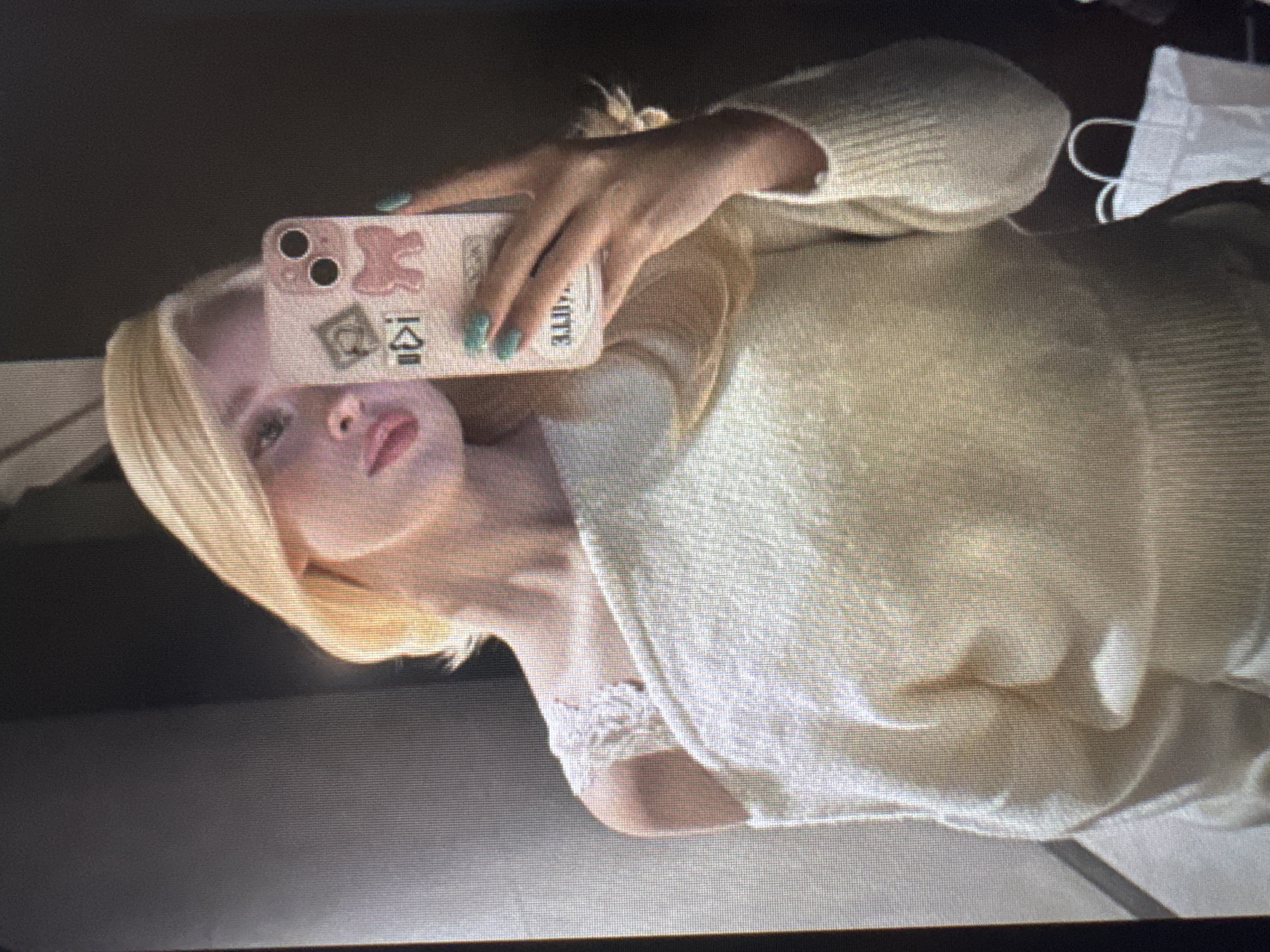 A sideways, high-angle selfie depicts a young woman with blonde hair and fair skin. She holds a light pink or beige smartphone with her right hand, her fingers adorned with light teal nail polish. The phone case is decorated with various stickers, including a pink bear shape, a square with a green dollar sign, and another square with a stylized 'K' or Korean character. Partially visible text on the side of the phone case reads "ATTE" or "OTTE."
The woman's gaze is directed towards the camera, her lips slightly parted in a neutral expression. She is wearing a light-colored, possibly cream or off-white, knitted sweater or top. Beneath the sweater, a white, lace-trimmed undergarment, likely a bra or camisole, is visible.
The background appears to be an indistinct indoor setting with a dark surface. A white disposable face mask with ear loops is visible in the upper right corner of the frame. The image exhibits prominent horizontal scan lines and a grainy texture, suggesting it may be a photograph of a digital display or screen.