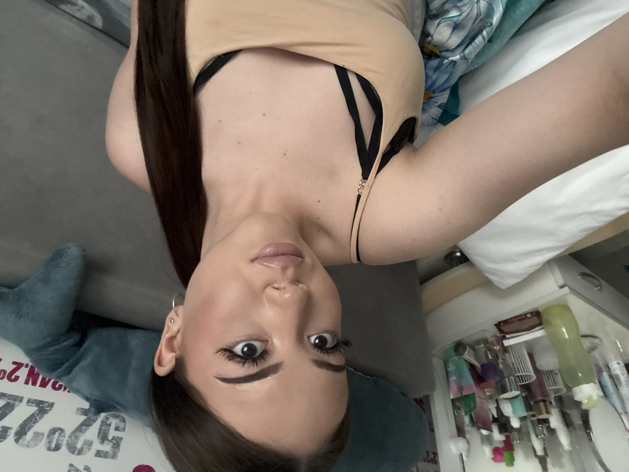 A person with fair skin, dark hair, and visible makeup lies on a bed. They are wearing a tan top with black straps. The bed has white and floral-patterned bedding. A blue plush object is also visible on the bed. A partially visible shelf cluttered with various containers and bottles is to the side. The location is unknown and cannot be determined from the provided image. It is not possible to tell from the image if this is Naberezhnye Chelny, Russia.