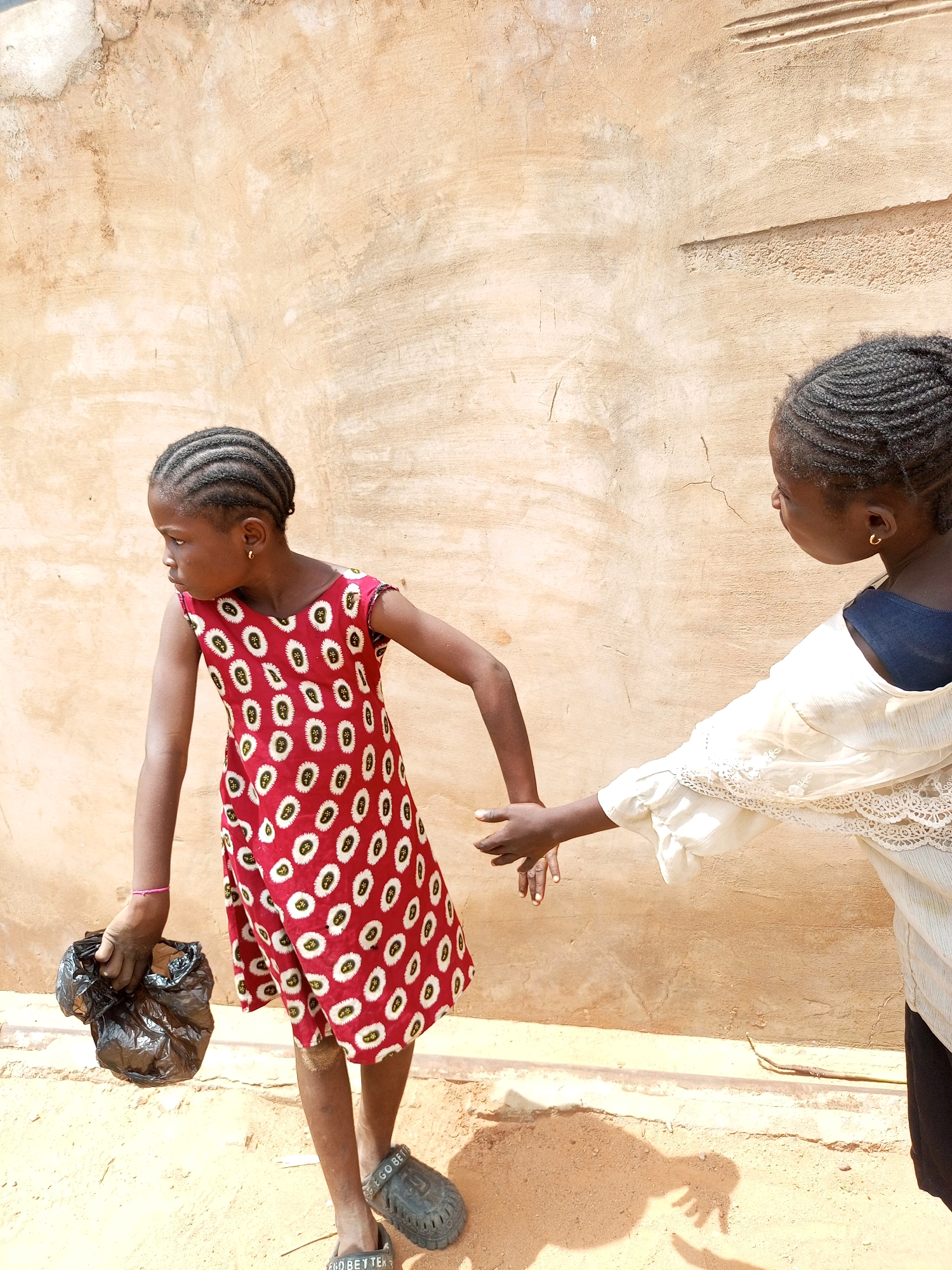 Two young girls are standing in front of a textured, tan-colored wall in Tempure, Nigeria. The girl on the left, facing away from the camera and looking to her left, wears a red dress with a white and yellow circular pattern. She is holding a black plastic bag in her left hand, which appears to contain some sort of contents. She has her hair braided in tight cornrows and is wearing dark, possibly black, clogs on her feet. A thin pink bracelet is visible on her left wrist. The girl on the right is partially visible, facing the girl on the left and extending her right hand towards her, palm open. She is wearing a light-colored, possibly off-white, garment with intricate lace trim around the shoulders and sleeves. A dark blue or black undershirt is visible beneath her garment. The ground beneath them is sandy or dirt. The wall behind them exhibits some cracks and textured imperfections. The overall lighting suggests it is daytime, possibly in the afternoon, casting shadows on the ground.