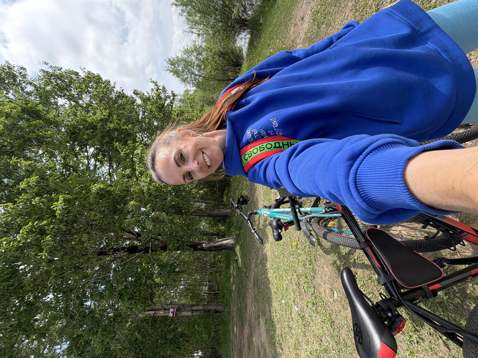 A young woman is taking a selfie outdoors. She's smiling at the camera and wearing a blue hoodie with lettering in what appears to be Cyrillic script, as well as a green and red striped arm band that also has Cyrillic writing. Two bicycles are visible; one is a blue mountain bike and the other is a black and red bicycle that has a child seat. The setting is a park or wooded area with green trees and grass. The location is believed to be Birobidzhan, Russia based on the Cyrillic lettering.