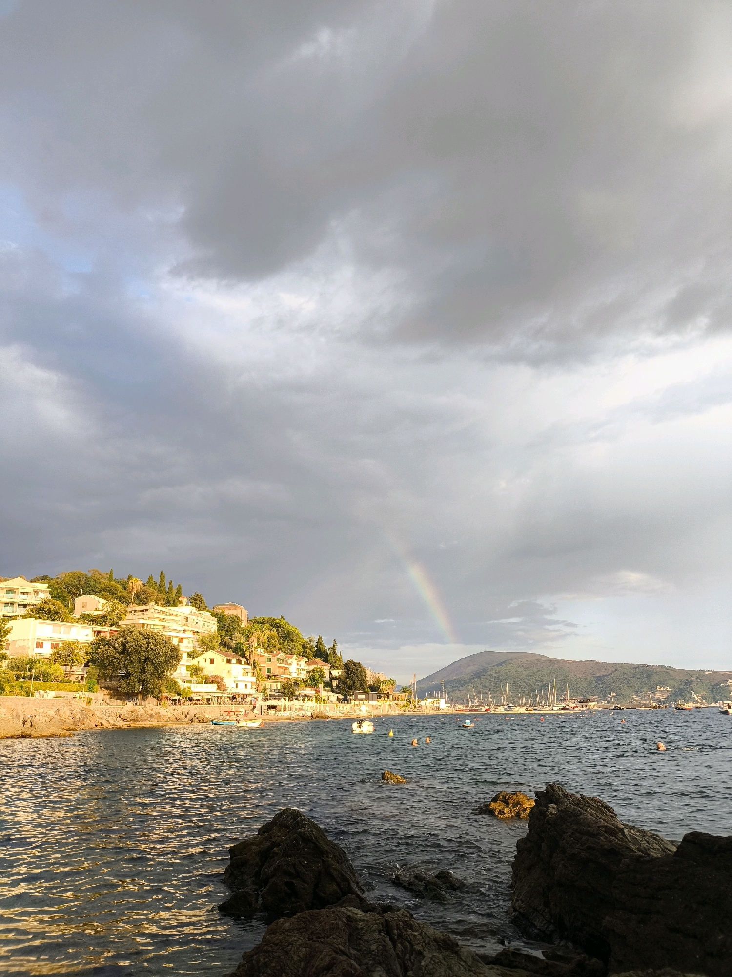 The image depicts a coastal scene with a town built on a hillside overlooking a body of water. The sky is filled with dramatic, dark clouds, with a faint rainbow arching across the horizon. The town's buildings are white and yellow, interspersed with green trees and cypress trees. Along the water's edge, there is a promenade with some restaurants and cafes. Several boats are moored in the harbor, and some people are swimming in the sea. In the foreground, there are dark, rocky outcrops. The overall impression is of a moody, atmospheric landscape with a touch of natural beauty. The location is identified as Herceg Novi, Montenegro.