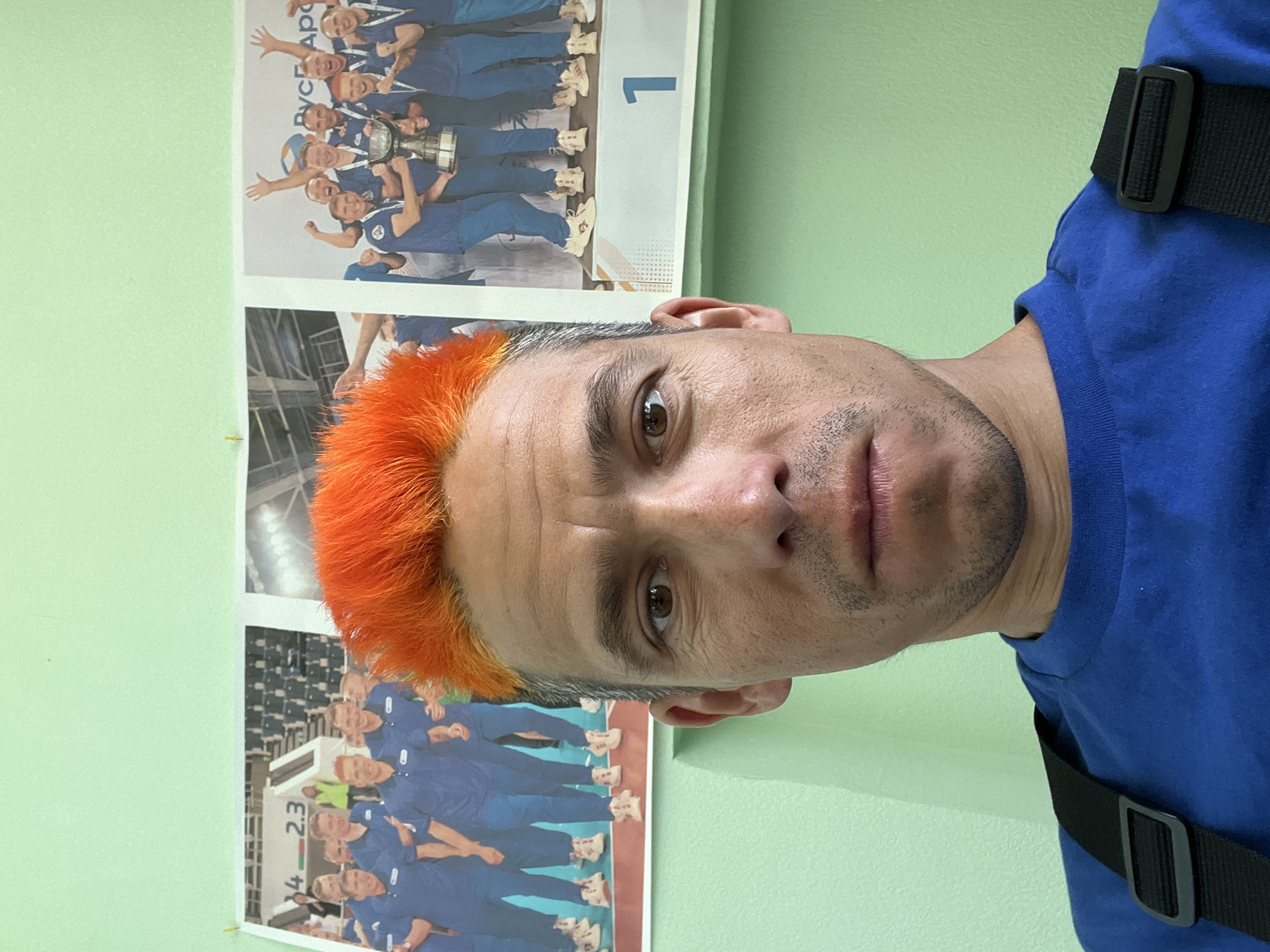 A medium shot shows a man with short, dyed orange hair. He is wearing a blue t-shirt and what appears to be a black strap or harness over his shoulder. The man is looking directly at the camera with a neutral expression.
Behind him on a light green wall are three printed posters featuring images of a sports team wearing blue uniforms. The top poster shows the team holding a trophy with the text "РУСГ Аро" and the number "1". The second poster shows the team indoors in what appears to be an arena, and the bottom poster shows the team grouped together on a stage, with partial text visible: "4-2.3".
Without further information or context, it is difficult to confirm the location in Chaikovsky, Russia. However, given the text "РУСГ Аро" (which could be an abbreviation for a Russian organization) and the team's uniform and general appearance, it is possible that the images depict a sports team from the city or region.