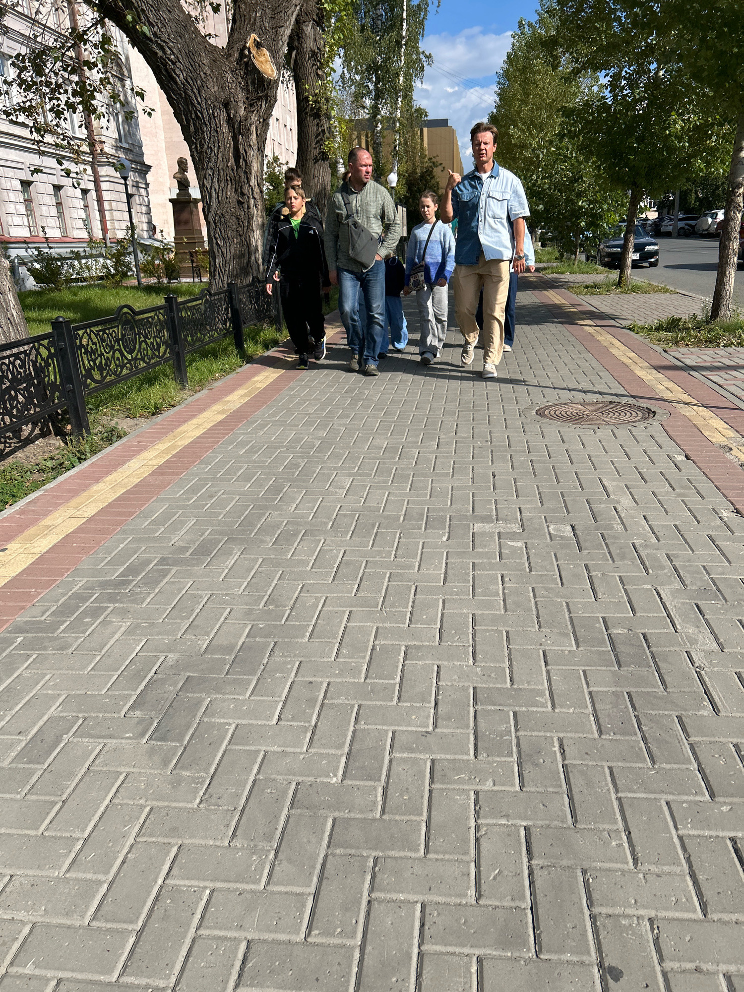 A group of five people, including two adults and three children, are walking along a paved sidewalk on a sunny day. The sidewalk is made of grey rectangular pavers arranged in a herringbone pattern, with a red and yellow border. On the left side of the sidewalk, there is a black metal fence, a grassy area, and a large tree. Beyond the fence, a building with classical architecture is visible, as well as a bust on a pedestal. On the right side of the sidewalk, there are more trees and a street with parked cars and a road. The people appear to be casually dressed. The man in the front right is wearing a light blue denim shirt and khaki pants, and is pointing to his left. The man to his left is wearing a green jacket and blue jeans. The children are walking in front of the adults. The scene suggests a leisurely stroll in an urban environment in Tomsk, Russia.