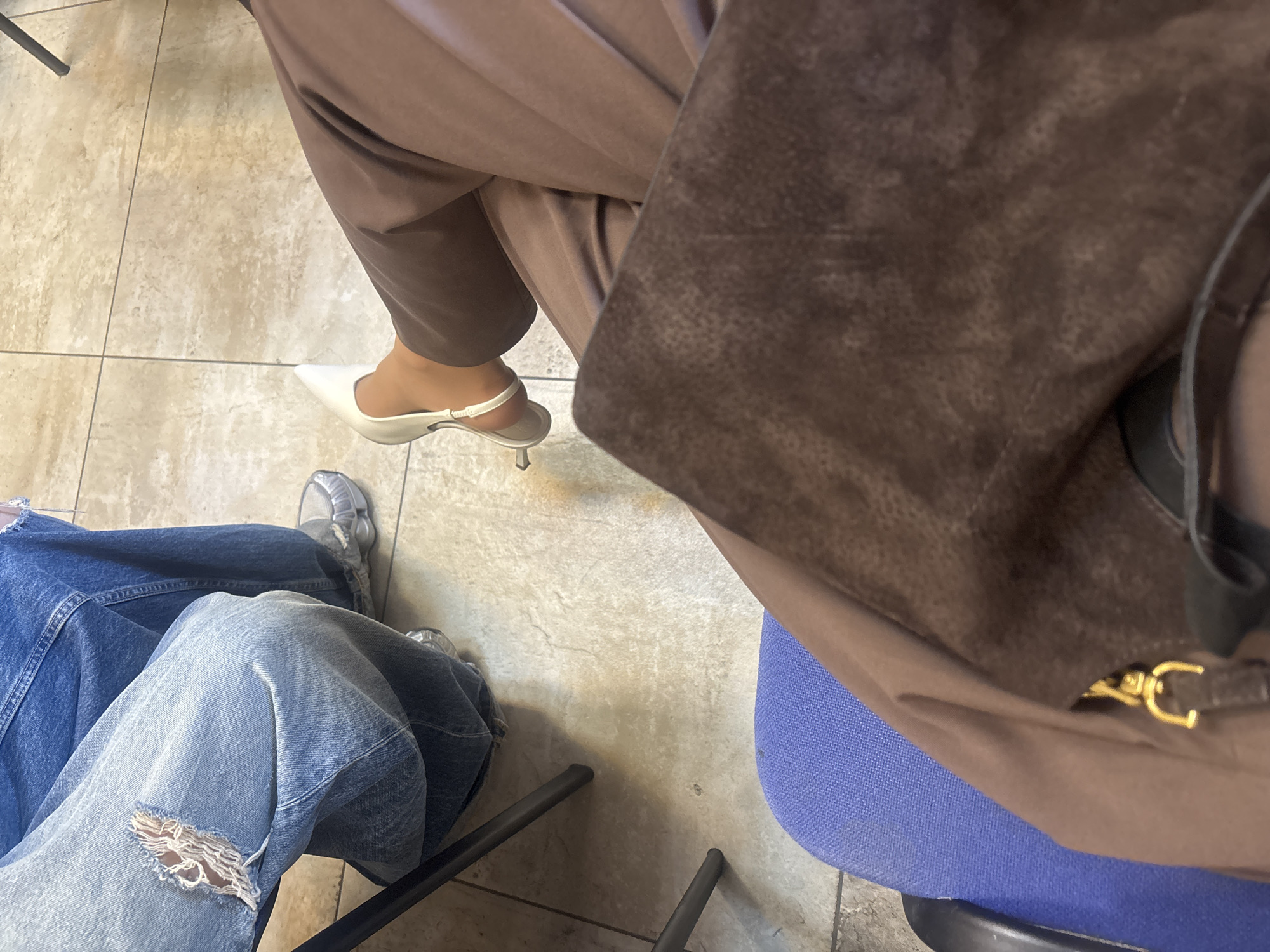 The image shows two people sitting in chairs, likely indoors due to the tile floor. One person is wearing tan pants and white heeled shoes, and their brown bag is partially visible. The other person is wearing blue jeans with a ripped knee and gray sneakers. A blue chair is also visible in the image. The specific location in Yerevan, Armenia, cannot be determined from the given visual information.