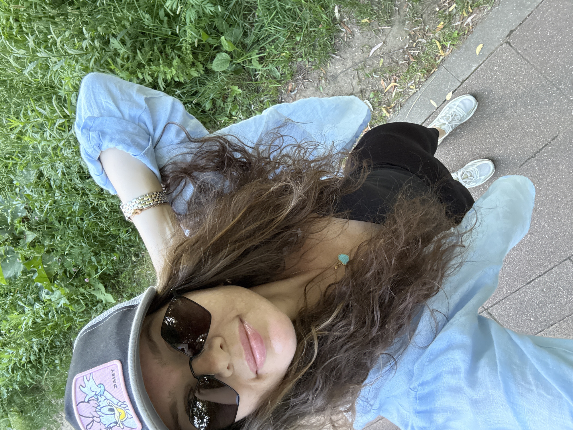 The image shows a woman lying on her back in a grassy area next to a paved walkway. She is wearing a light blue shirt, a black skirt, and white sneakers. She has long, curly brown hair and is wearing sunglasses and a baseball cap. A small turquoise pendant necklace is visible. The scene appears to be outdoors, possibly in a park, with green grass and trees in the background. The lighting suggests it is daytime, with mild weather. The woman is looking up and taking a selfie, with a relaxed and content expression. A patch on her cap has the word "DAISY" visible on it. The overall impression is one of leisure and enjoying a pleasant day outdoors.