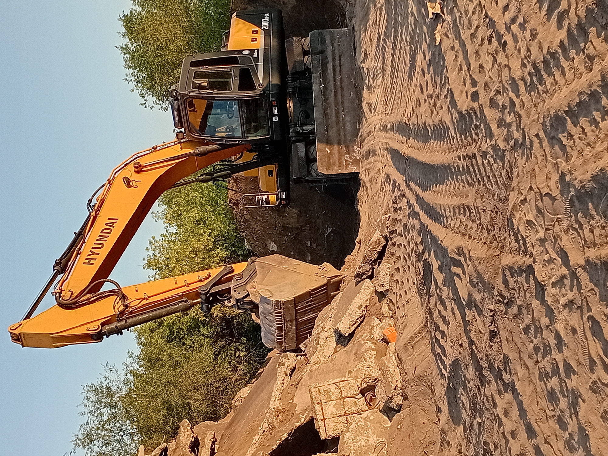 An orange Hyundai excavator is positioned on a construction site in Semey, Kazakhstan. The excavator is actively engaged in demolition or excavation, with its bucket digging into a pile of concrete debris and earth. The surrounding environment consists of sandy ground with visible tire tracks from the machinery, and a backdrop of green trees under a clear sky. The scene suggests ongoing construction or demolition work, typical of a developing or redeveloping urban area.