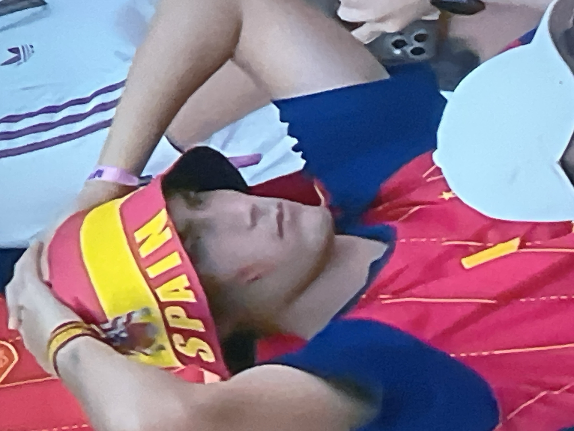 The image shows a close-up of a young person wearing a red and yellow bucket hat that says "SPAIN" in yellow letters. The hat has a red band with yellow stripes. The person is also wearing a red jersey with blue sleeves and a yellow stripe. Their hands are on top of the hat. In the background, a person in a white shirt with purple stripes and an Adidas logo is visible, along with a white baseball cap and a portion of a cell phone. The scene appears to be outdoors, possibly at a sporting event or gathering. The provided context that the image is located in Jos, Nigeria, is not visually verifiable from the image itself, as there are no discernible landmarks or environmental clues specific to that city.