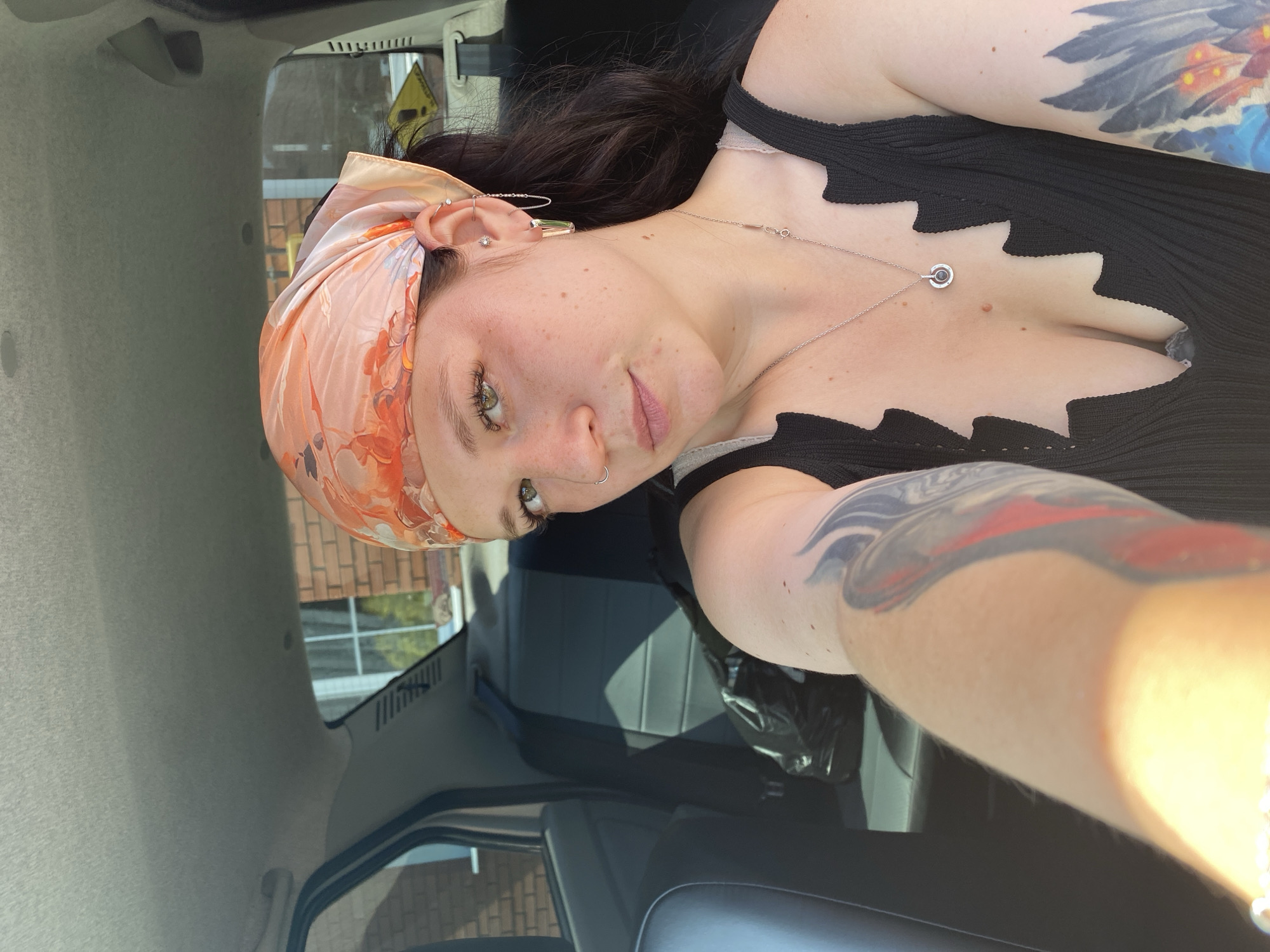In the city of Fokino, Russia, a woman with dark hair and a headscarf with an orange floral pattern is sitting in the passenger seat of a car. She is looking at the camera, and her left arm is extended outwards, holding the camera for a selfie. She has tattoos on both her arms and is wearing a black top with a unique scalloped neckline and a silver necklace. The interior of the car, including the back seats and headliner, is visible behind her. The scene is illuminated by sunlight.