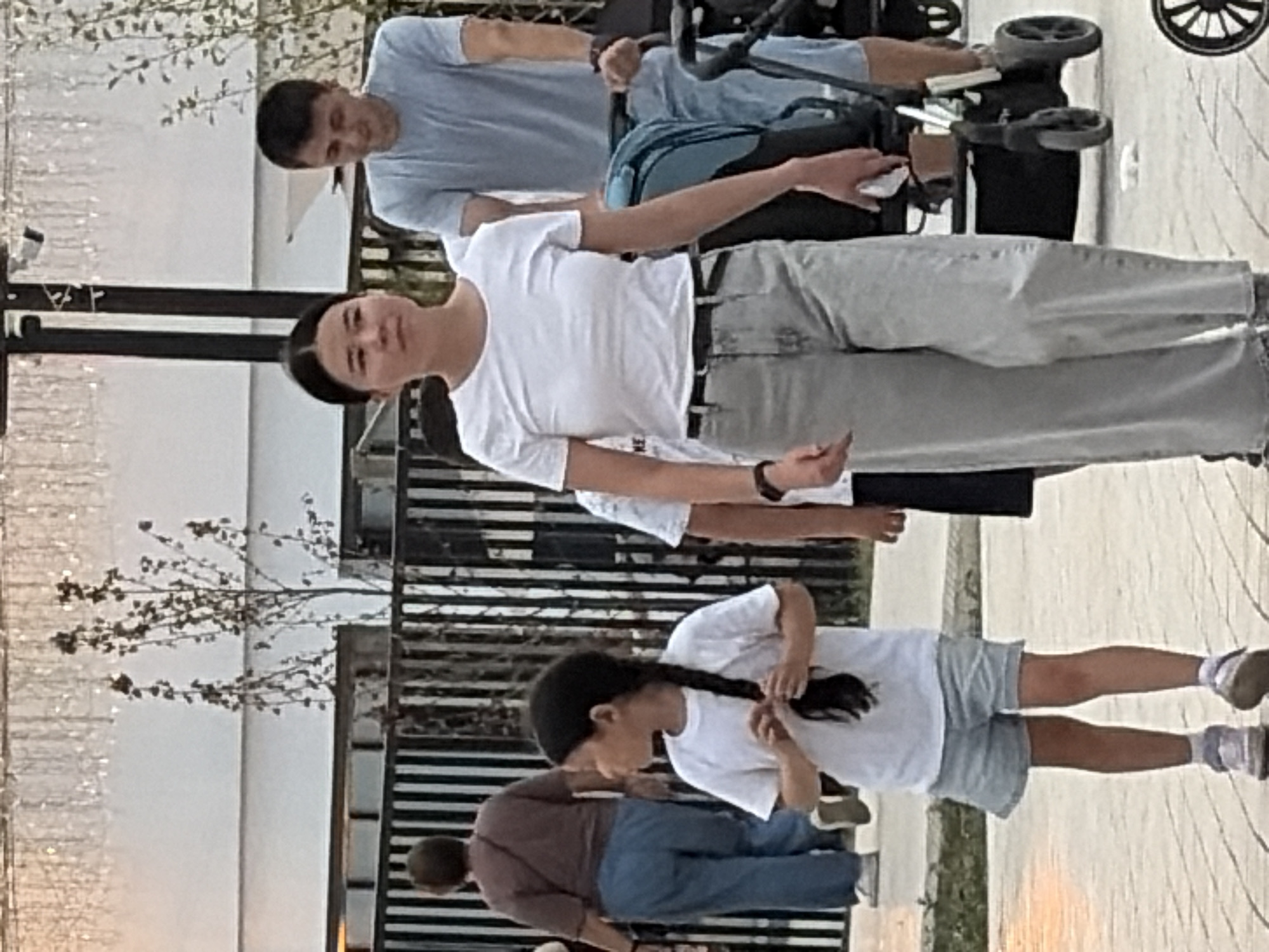A vertical, full-shot, eye-level photograph shows a group of people outdoors. The scene appears to be a paved area with a black metal fence in the background. A young woman in a white t-shirt and gray pants is in the foreground, facing right, with her arm outstretched. Behind her, a young girl in a white t-shirt and gray shorts is also facing right, with her hair in a braid. She appears to be touching her hair. In the background, two men are visible. One, wearing a light blue t-shirt and jeans, is looking down towards a stroller. The other, wearing a dark shirt, is partially obscured. The time of day is unclear, but the lighting suggests it might be daytime. The weather is not discernible. No visible text is present in the image.
