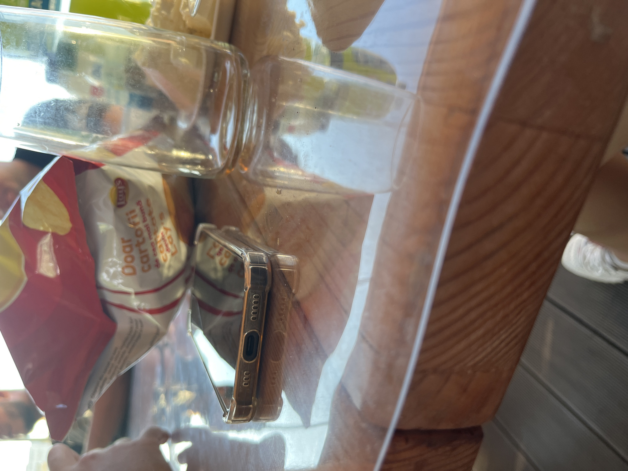 The image depicts a close-up, eye-level, outdoor scene with a focus on a table surface. On the table, a bag of Lay's potato chips is partially opened, with text in Romanian visible, including "Doar cartofi". A clear glass bottle, appearing to be a beverage container, is placed next to the chip bag, and is empty. A smartphone, with its charging port visible, rests on the table, its screen reflecting the surroundings. The table appears to be made of wood with a glossy, possibly plastic, overlay. In the background, out of focus, a person is partially visible. The overall setting suggests a casual outdoor gathering. The scene is set in Kazanlak, Bulgaria.