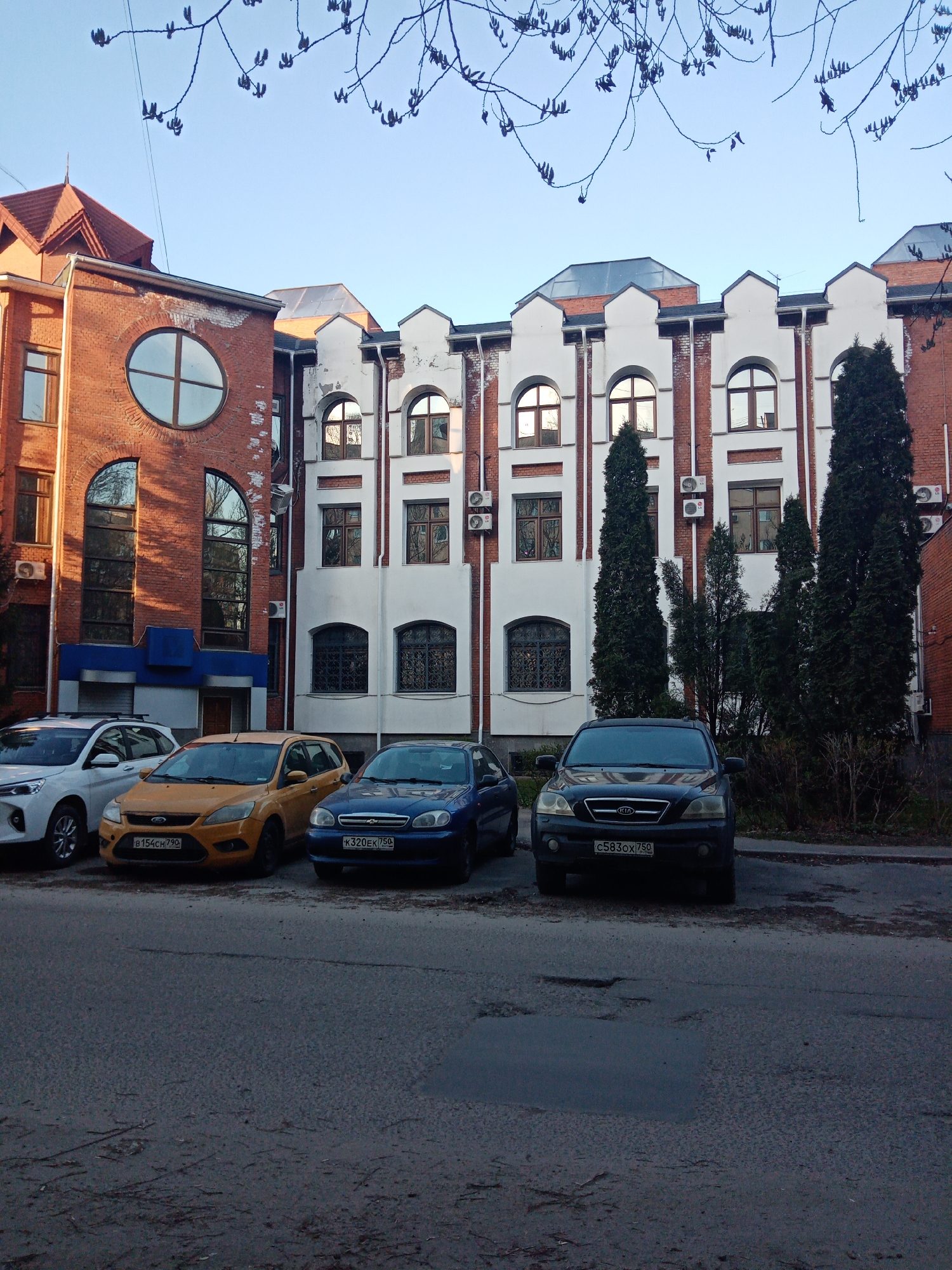 The image captures an urban street scene in Elektrostal, Russia, likely during late winter or early spring, as evidenced by the bare tree branches with visible buds against a clear blue sky. The light suggests either early morning or late afternoon.
In the foreground, an asphalt road displays uneven textures and some darker, possibly wet, patches. Four cars are parked side-by-side, facing the viewer. From left to right, these are a white SUV (resembling a Hyundai Creta), a yellow Ford Focus hatchback bearing the Russian license plate "B154CH790", a blue Chevrolet Lanos sedan with the license plate "K320EK750", and a dark Kia Sorento SUV displaying the license plate "C583OX750". The license plates indicate registration in Russian regions (790 and 750 are associated with Moscow and Moscow Oblast).
Behind the parked vehicles stand two distinct, adjoining buildings. The structure on the left is a multi-story, red-brick building with a prominent large circular window on its upper floor and tall, arched windows below, reflecting the sky and surrounding environment. It features a striking, multifaceted reddish-brown roof. Connected to its right, the second building presents a different aesthetic, characterized by a facade with white decorative elements over red brick, featuring numerous arched windows on various levels. Several tall, slender evergreen trees are planted directly in front of this building, partially obscuring some windows. Multiple outdoor air conditioning units are visible on its facade. No people are present, and the overall scene conveys a calm, everyday urban environment.