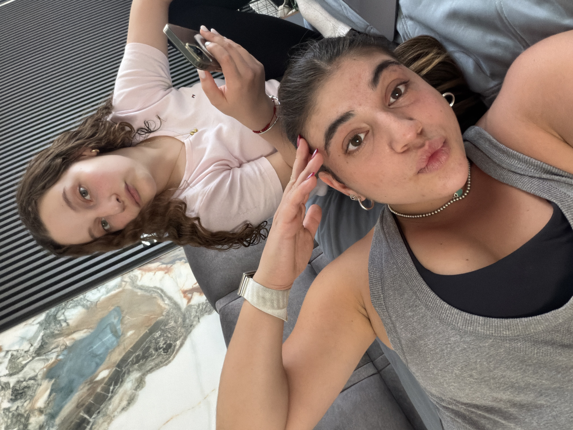 Two young women are lying on a grey upholstered piece of furniture, possibly a sofa or chaise lounge. The woman in the foreground, dressed in a grey tank top over a black sports bra, is closer to the camera and making a kissing face while holding her hand up near her forehead. She is wearing a silver choker necklace, hoop earrings, and a white smartwatch. The other woman, dressed in a pink t-shirt, is lying behind her and holding a gold mobile phone. The background includes a striped wall and a marble floor or table surface. The location in the city of Puebla, Mexico, is not discernible from the image.