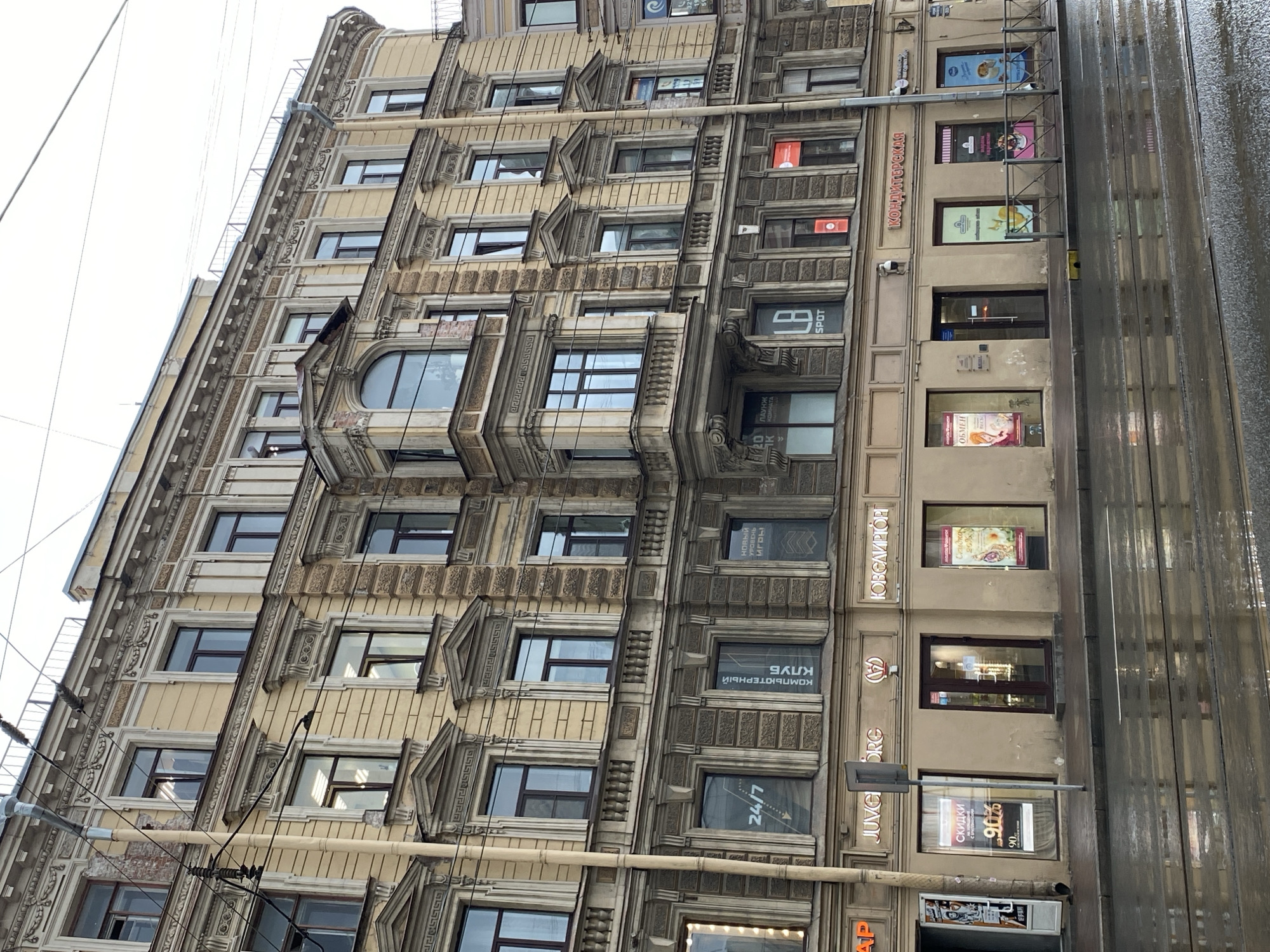 The image shows a multi-story building with an ornate facade, typical of Saint Petersburg, Russia. The building features classical architectural elements such as columns, cornices, and decorative moldings. The facade is painted in a pale yellow color with darker accents.
At street level, there are several storefronts. Signs indicate various businesses, including "JUVELIORG" (jewelry), "КОМПЬЮТЕРНЫЙ КЛУБ" (computer club), and "КОНДИТЕРСКАЯ" (confectionery). Some of the windows display advertisements for discounts and products.
The street in front of the building is wet, suggesting recent rain, and tram tracks are visible. The sky is overcast, contributing to a somewhat muted lighting in the scene. There are no people visible in the image. Electrical wires are strung across the upper part of the building and the sky. The architectural style and signage strongly indicate this is a commercial street in Saint Petersburg.