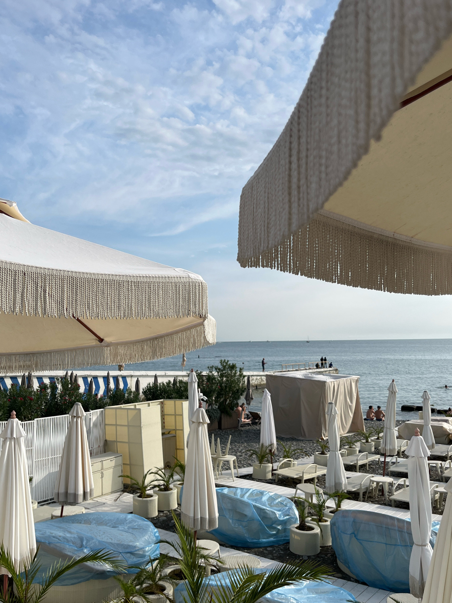 The image captures a view of a beach resort in Sochi, Russia, on a partly cloudy day. In the foreground, several white beach umbrellas with decorative fringes are visible, with some furniture covered in blue tarpaulin. Behind them, a white fence encloses a section of the resort with more umbrellas and seating. The middle ground shows the beach with loungers, some covered, and scattered umbrellas. In the background, the Black Sea stretches out, with a few people on a pier and others in the water. The sky is a light blue with scattered white clouds. The overall impression is a peaceful, somewhat quiet beach setting, possibly before the main tourist season or in the late afternoon.