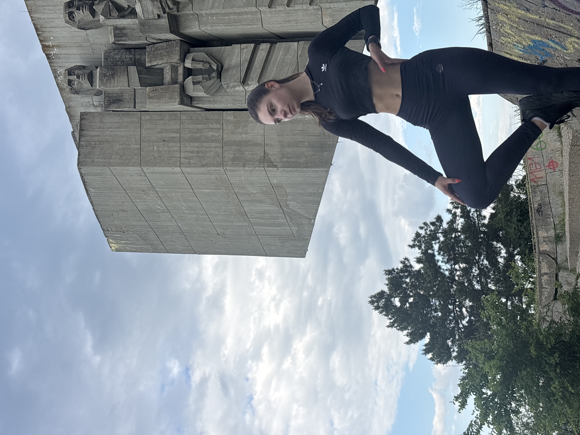 The image captures a young woman, likely in her late teens or early twenties, posing in front of the Monument of the Bulgarian-Soviet Friendship in Varna, Bulgaria. The monument, a large concrete structure with abstract sculptural elements, dominates the left side of the frame. The woman is wearing black athletic attire consisting of a long-sleeved crop top and leggings, and black sneakers. She has long brown hair, and her pose is casual with one hand on her hip. The sky is partially cloudy, suggesting it's daytime. To the right, there's greenery, including a tree, adding a natural element to the scene. The overall impression is that the woman is engaging in a photo shoot at this landmark.