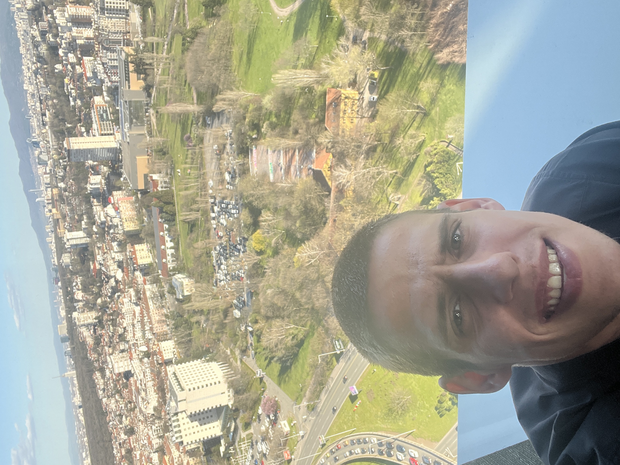 A low-angle, selfie-style photograph shows a man smiling at the camera, with an expansive cityscape visible in the background. The city is Sofia, Bulgaria, discernible from the distinct urban layout. The man has short hair and is wearing a dark-colored top. The cityscape includes a mix of low and high-rise buildings, dense urban development, and green spaces. The urban landscape seems to stretch towards distant mountains under a clear sky. A park area with sparse trees and winding pathways can be seen between the buildings. The photo seems to be taken from a high vantage point, probably a rooftop or a tall building.