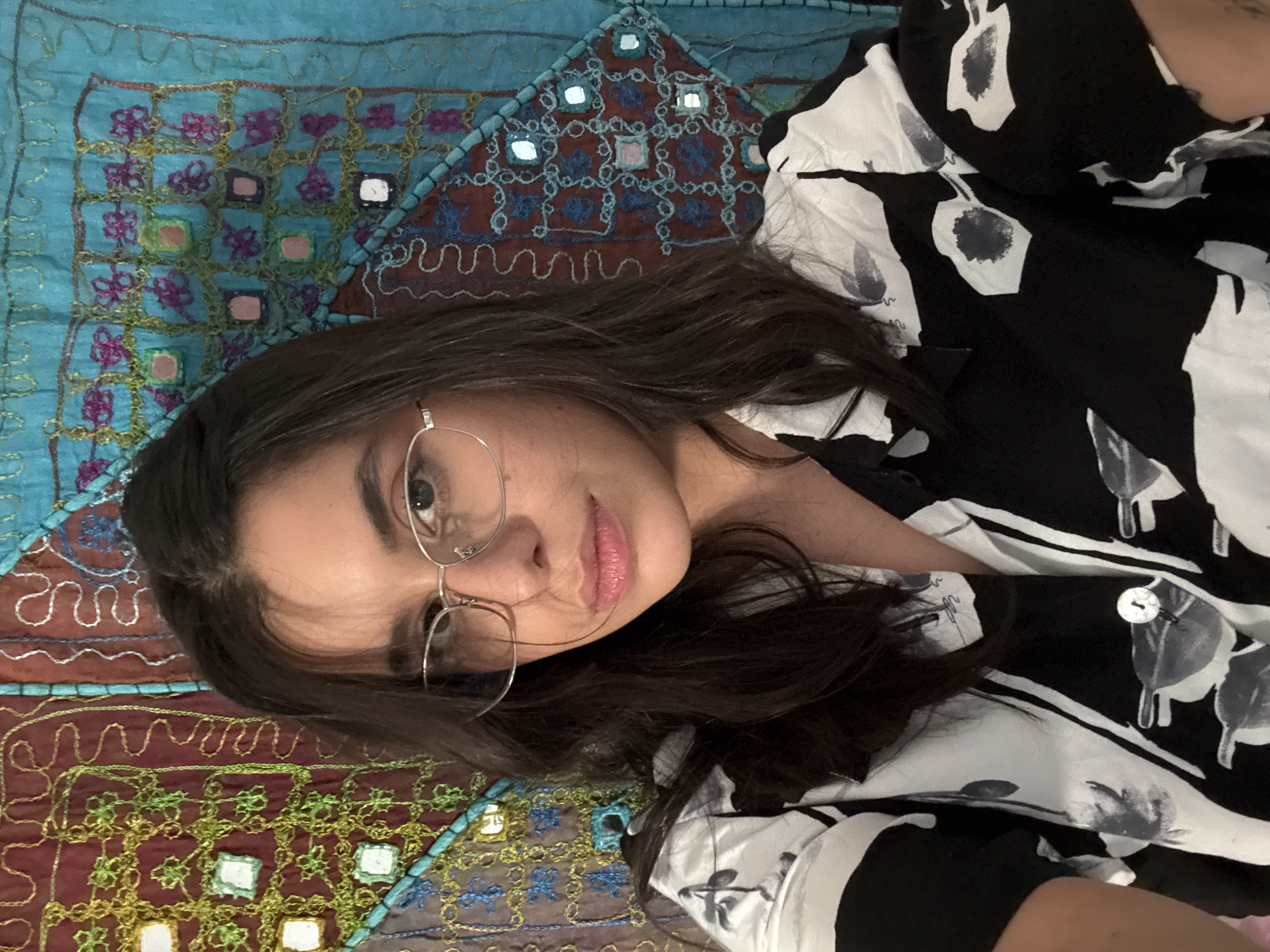 In an indoor selfie, a young woman with long, dark hair and wearing thin, wire-rimmed glasses looks directly at the camera. Her lips are slightly parted, and she has subtle makeup on. She is wearing a black and white collared shirt with a floral pattern. The background consists of a colorful tapestry with intricate embroidery and small mirror-like accents. The tapestry features a mix of blues, greens, reds, and purples. The lighting appears to be natural, coming from the front and slightly from the left. The scene is static, with no discernible action taking place. The overall impression is one of a casual, personal portrait. There is no information in the image to indicate the location in Las Rozas de Madrid, Spain.