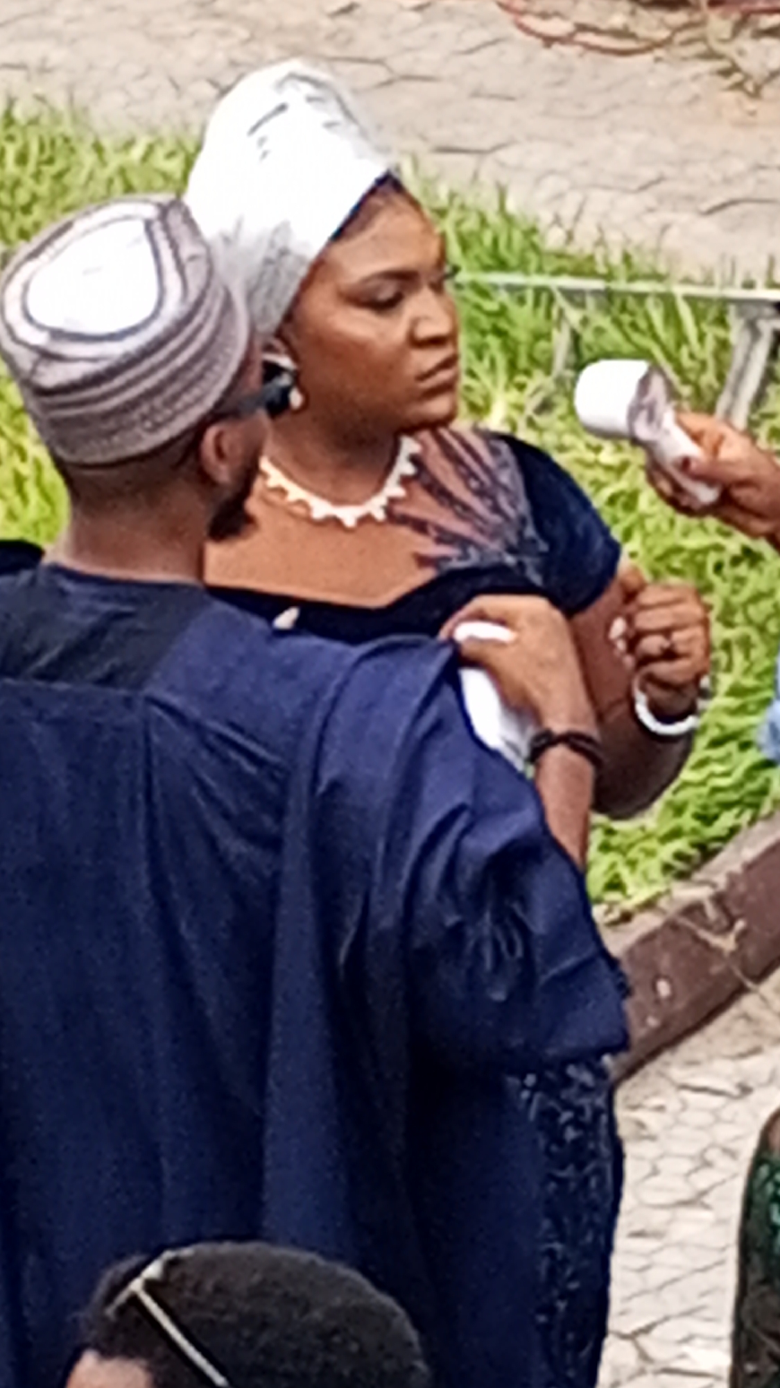 A woman in a dark blue velvet dress, a white necklace, and a white headwrap is being interviewed with a microphone. She is the focal point of the image. To her left is a man with a dark-striped hat, wearing a dark blue robe-like garment, facing away from the camera. Another person is to her right holding the microphone. The background includes greenery and a blurred pathway. The location is speculated to be in the city of Lokoja, Nigeria, without further evidence.