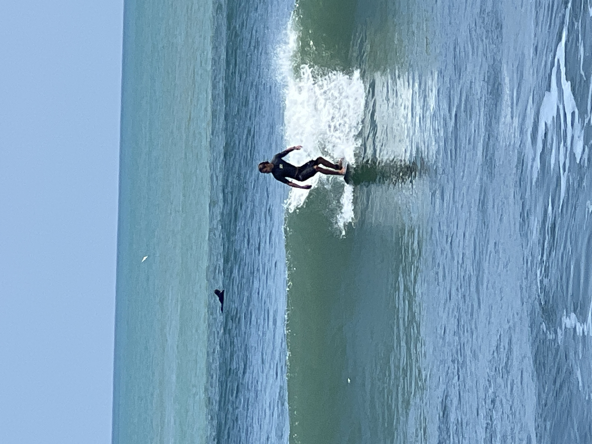 A male surfer wearing a black wetsuit is riding a wave on a surfboard. He is positioned in the middle of the frame, with the wave breaking around him. The ocean is a clear turquoise color and the sky is a bright blue. In the distance, another person is lying on a surfboard in the water. The image appears to be taken on a sunny day, with good visibility. The scene is consistent with a beach location, such as the popular surf spot of Kuta, Indonesia.