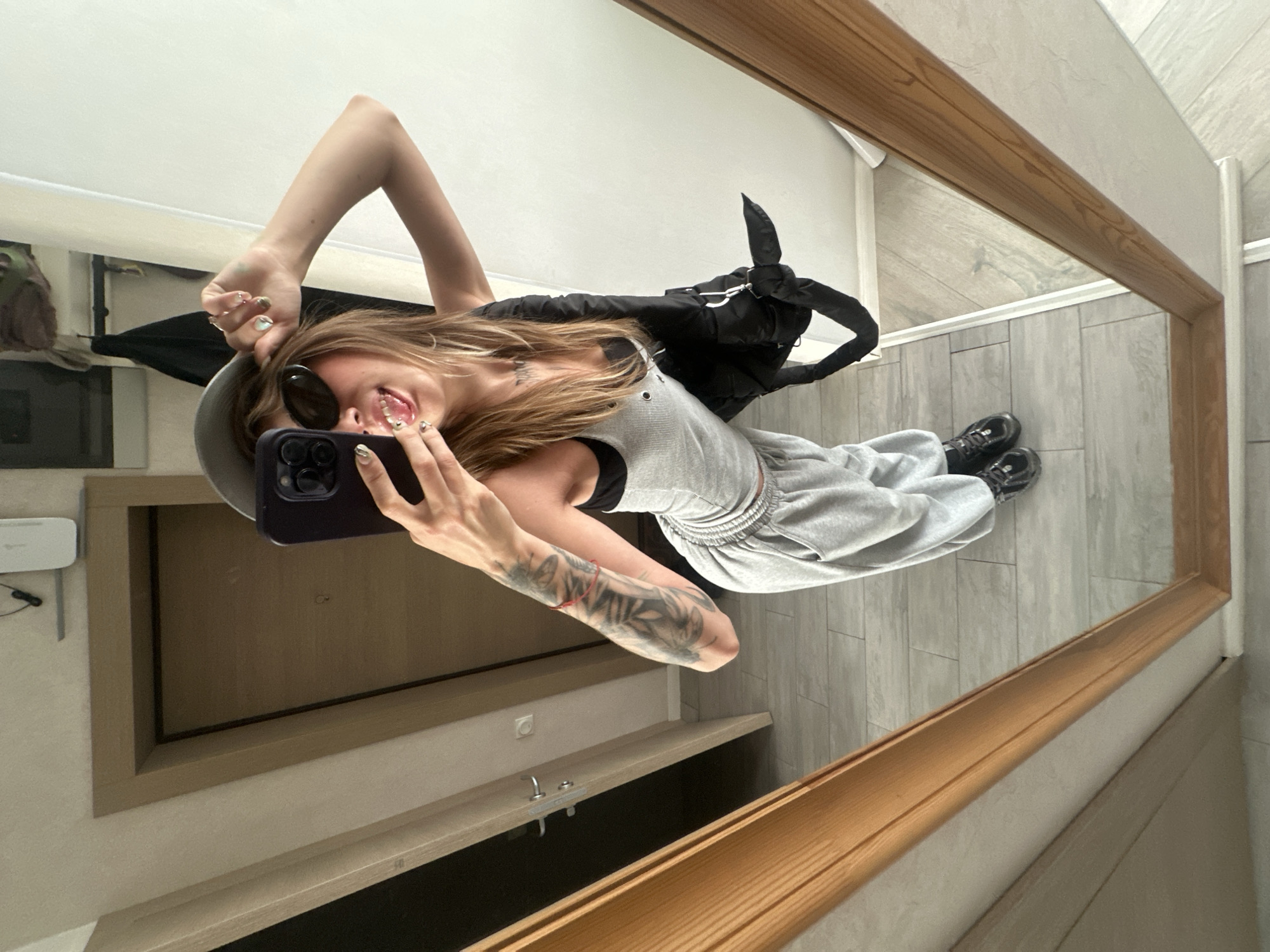 A young woman with blonde hair, wearing sunglasses and a grey tank top with grey sweatpants, poses for a selfie in a mirror. She is also wearing a grey baseball cap and carrying a black backpack over her shoulder. Her left arm is tattooed with floral designs. The background shows an interior hallway with a door and a wooden-framed mirror. The floor is tiled. There is no indication of the specific location within Moscow, Russia.
