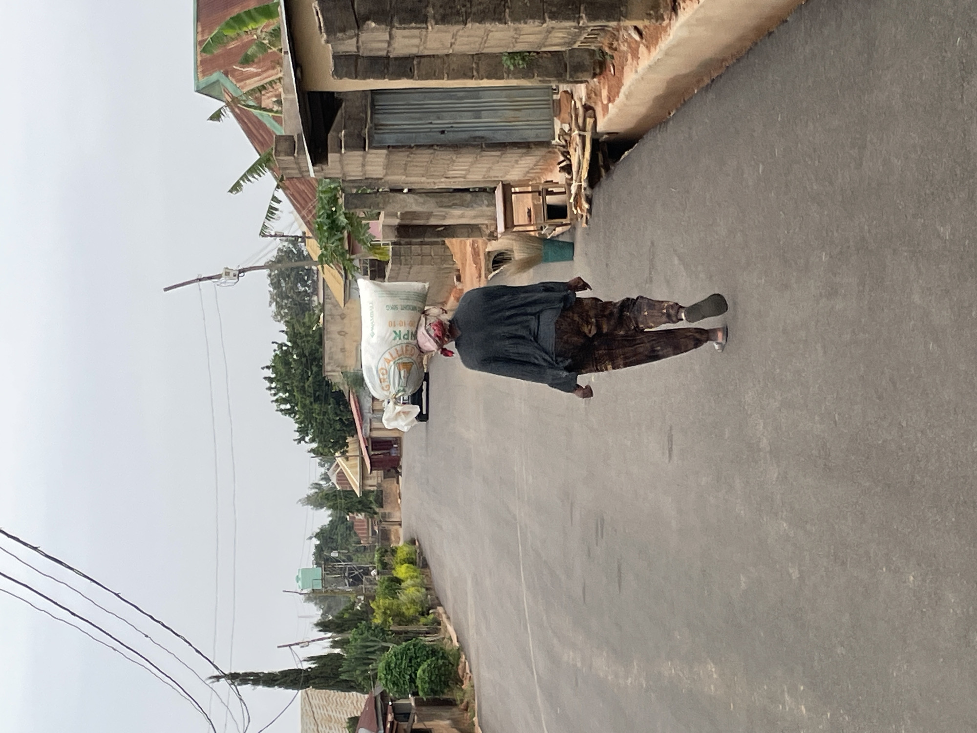 The image, taken in Jos, Nigeria, captures a person walking along a paved road, carrying a large white sack on their head. The individual is dressed in a dark long-sleeved top, patterned trousers, and sandals, with a reddish head covering visible. The white sack clearly displays the text "NPK 15-15-15 50KG", indicating it likely contains fertilizer.
The scene is an outdoor street environment with buildings lining one side of the road. These structures are made of concrete blocks and feature corrugated metal roofs, some with a green patina and others showing rust. On the opposite side of the road, and further into the background, there is abundant greenery, including trees, shrubs, and plants. Utility poles and electrical wires are visible overhead, particularly on the left side of the frame. In the distance, a light green water tank can be seen.
The sky is overcast or hazy, creating diffused lighting across the scene, suggesting daytime hours under cloudy conditions. The overall atmosphere is quiet and functional, with the person focused on transporting their load. The architectural styles, presence of utility lines, and method of carrying goods are consistent with a local street scene in a Nigerian town or city.
