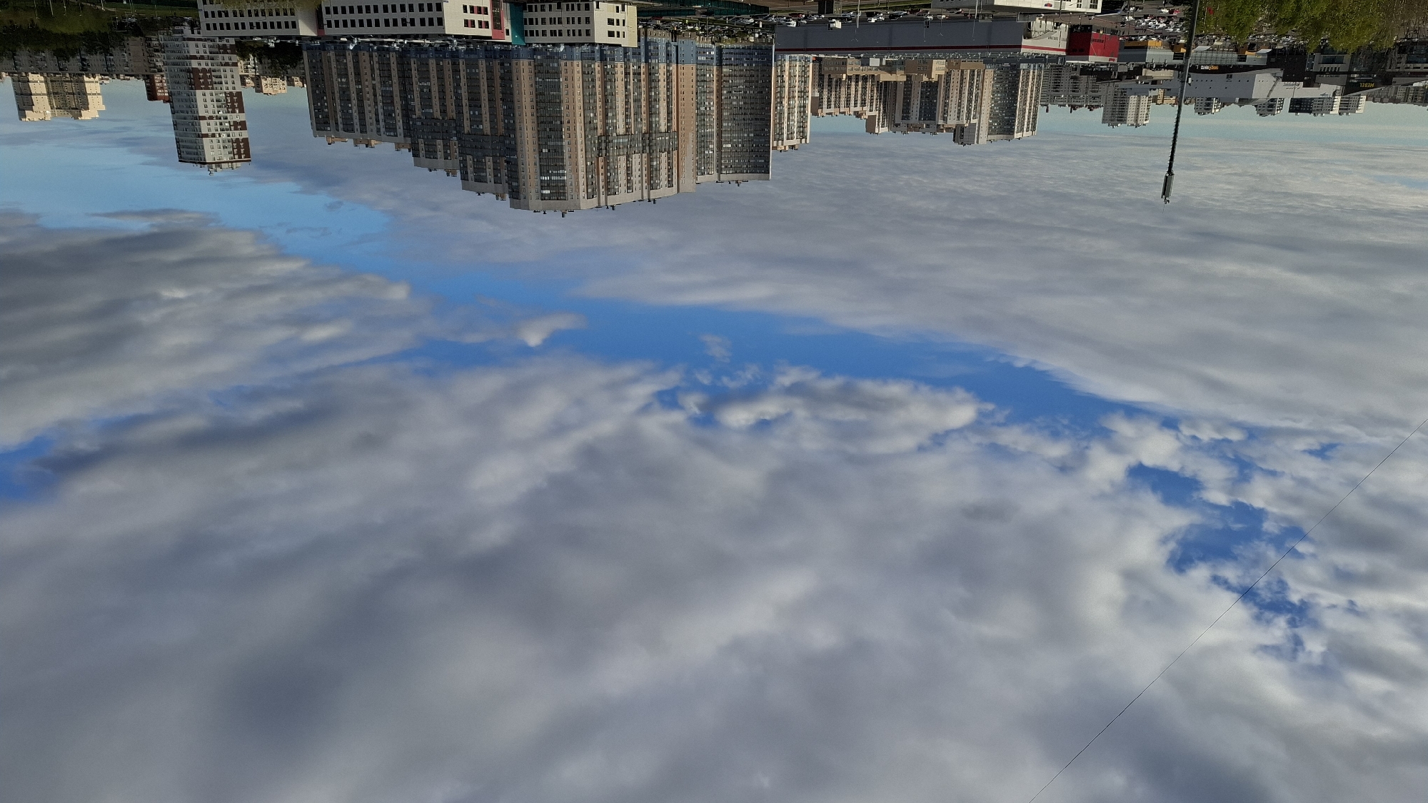 A high-angle shot captures a city landscape in Rakhya, Russia, with the scene inverted. The sky dominates the view, filled with a mix of gray and white clouds. A central patch of blue sky provides a break in the cloud cover. The city's reflection appears above the clouds, featuring modern apartment buildings, commercial structures, and greenery. A tall, thin pole stands out against the skyline.