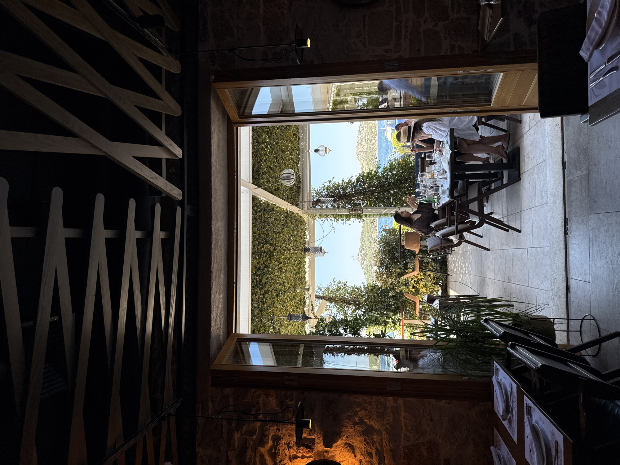 The image captures an indoor dining area of a restaurant with a view of the outdoors. The interior features stone walls and a dark, wooden lattice ceiling. In the foreground, tables are set with white napkins and cutlery, suggesting a meal in progress or preparation. Through an open doorway, an outdoor seating area is visible, covered with a green, leafy canopy. Several people are seated at tables in the outdoor area, engaged in dining and conversation. One woman wears a straw hat. Beyond the outdoor seating, a body of water with hills and islands in the background indicates a coastal location. The overall scene suggests a pleasant dining experience with a scenic view. The location is identified as Grad Šibenik, Croatia, a coastal city known for its historical sites and proximity to islands and the sea.