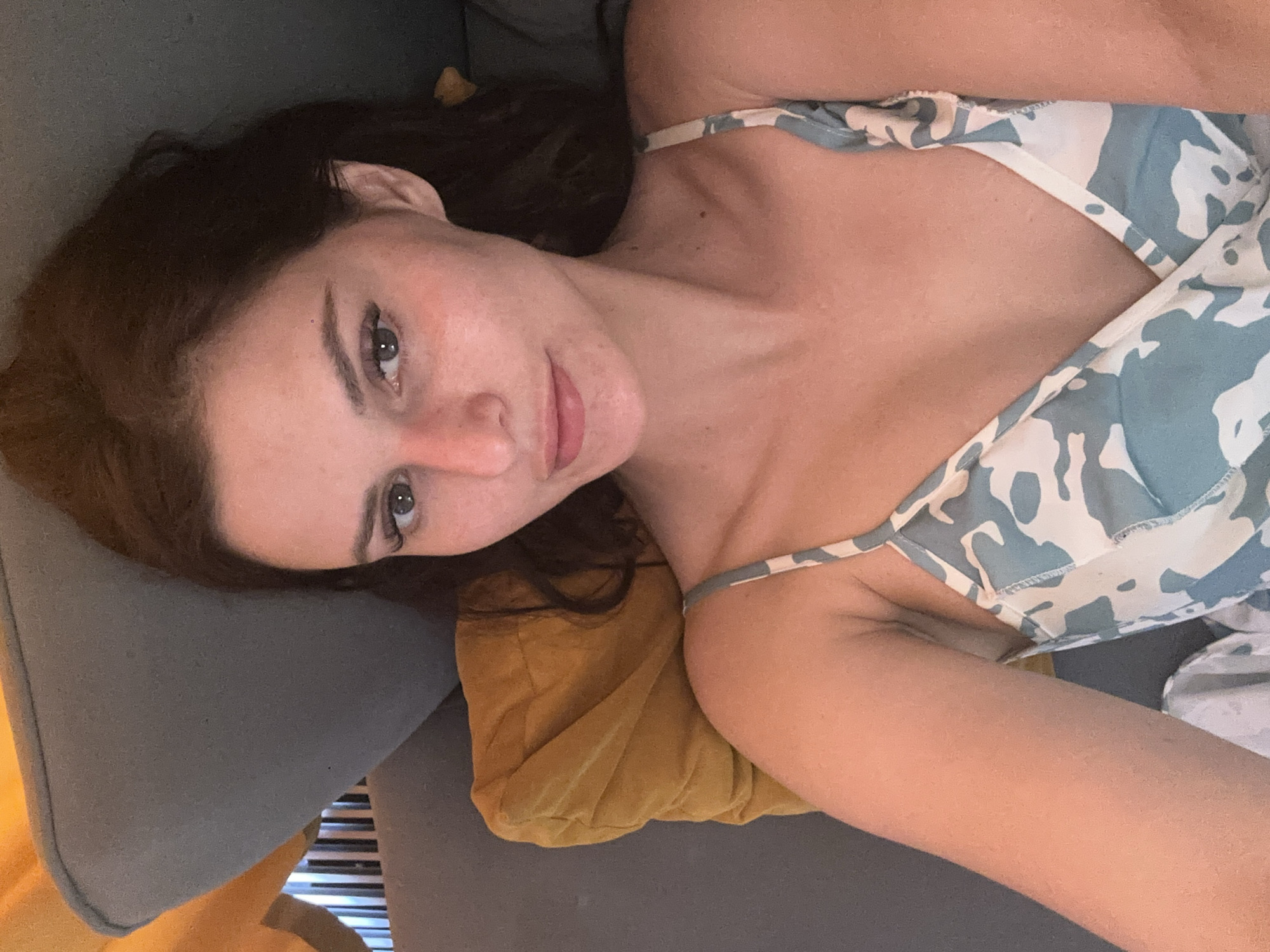 An unidentifiable woman, who appears to be of East European descent, is captured in a close-up, waist-up, selfie-style portrait, taken from a high angle. She is lying down, with her head tilted towards the camera and her body angled slightly to the left. The woman has dark brown hair, styled in loose waves, and fair skin with a smattering of freckles across her nose and cheeks. Her eyes are a light green hue, and her lips are painted a soft pink. She is wearing a light blue and white patterned camisole.
The woman is positioned on a gray couch, with a yellow pillow behind her head. To the left of the frame, a portion of a gray radiator can be seen. The background is a blurred mix of dark grays and hints of yellow, suggesting an indoor setting with dim lighting. No specific location within a city or country can be determined from the image.