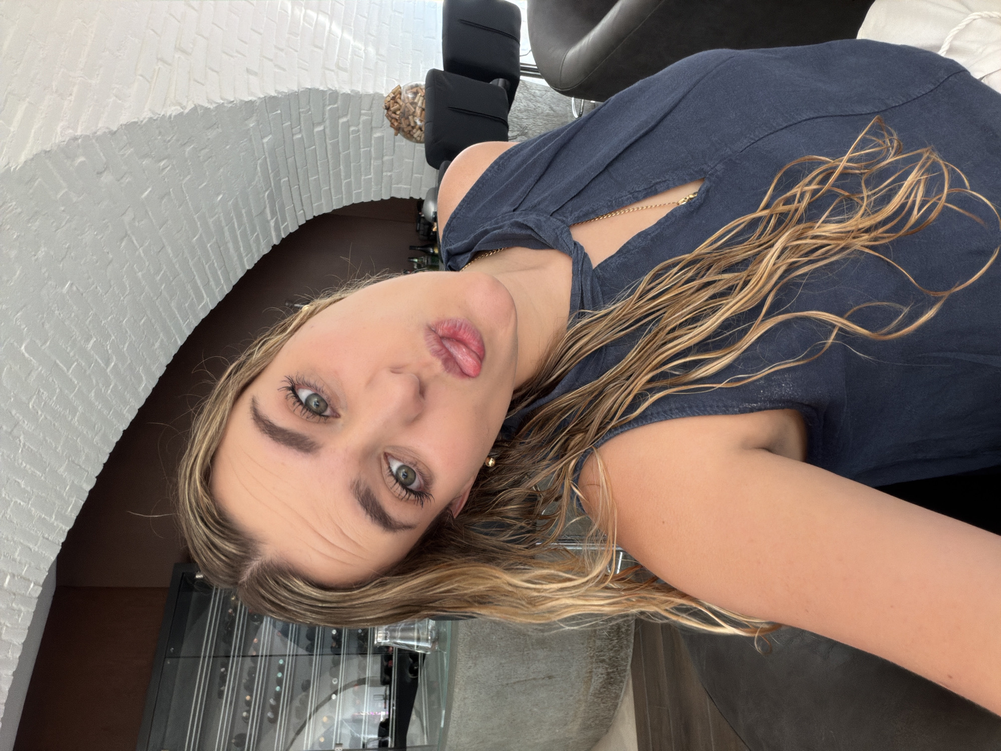 The image displays an adult female in a selfie, reportedly located in San Bernardino Tlaxcalancingo, Mexico. The subject has fair skin, long, wavy, blonde and light-brown hair, appearing wet, and green eyes. Her head is tilted to the left, and she exhibits a playful expression with puckered lips and a partially extended tongue. She is wearing a dark blue sleeveless top with a keyhole neckline, a thin gold-colored chain necklace, and a single spherical gold-colored earring on her visible right ear.
The background on the left side features a dark brown wall and a glass-fronted cabinet displaying multiple bottles, possibly wine. A clear glass decanter rests on a grey, textured surface. The right side of the background reveals a prominent architectural element: a white-painted brick arch. Beneath the arch, two black bar stools are visible, alongside a clear glass bowl filled with numerous small, brown, irregular solid objects. Dark-colored upholstered furniture, likely chairs, is partially visible in the lower left and right portions of the frame, suggesting an indoor seating area.
