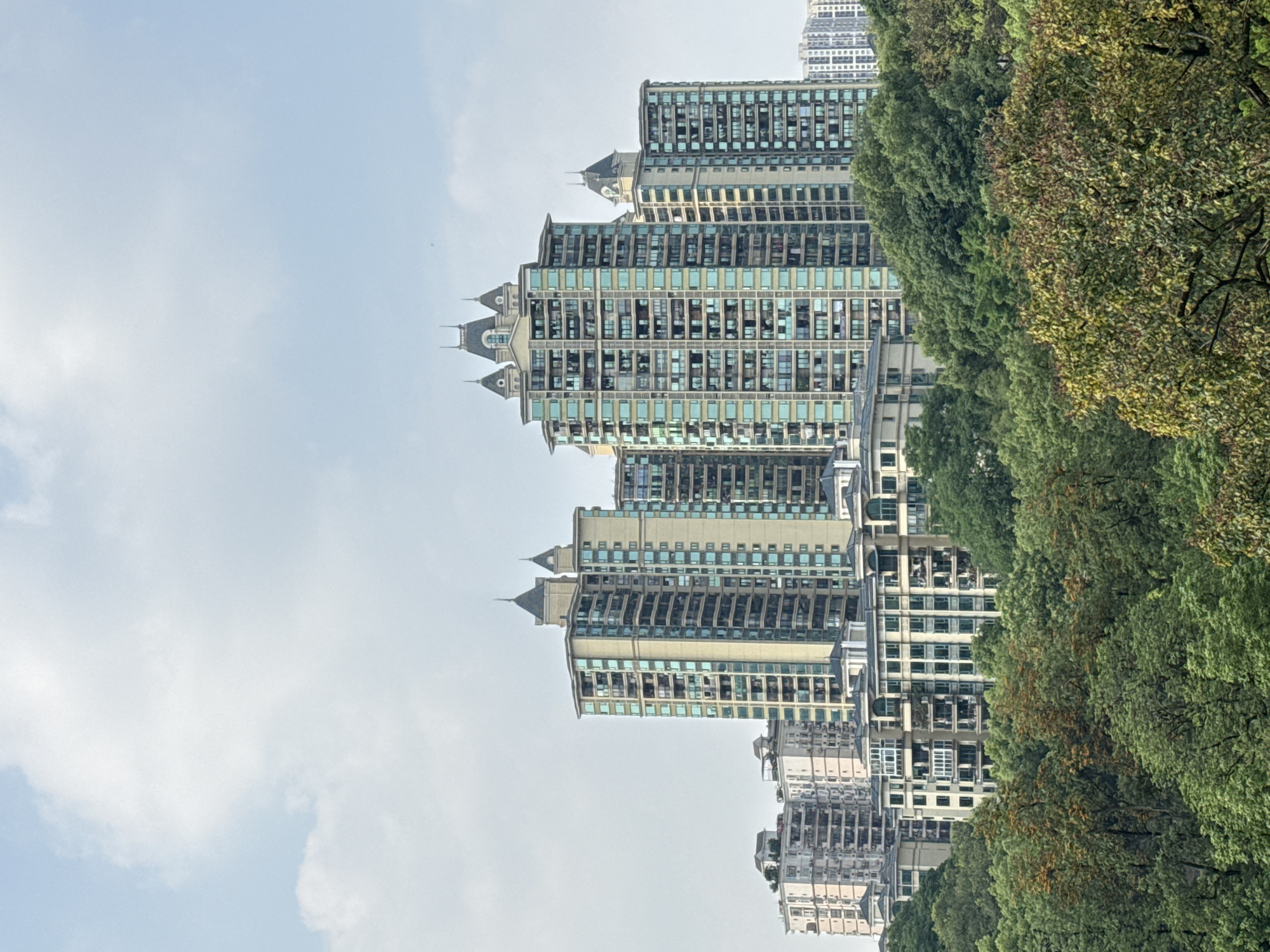 The image captures a view in Jiulongpo District, Chongqing, China, showcasing high-rise residential buildings adorned with distinct architectural details like pointed turrets. These structures are partially obscured by dense, green trees with hints of autumnal coloration. The sky appears overcast, with a mix of light blue and white clouds. The image suggests an urban setting juxtaposed with natural elements.