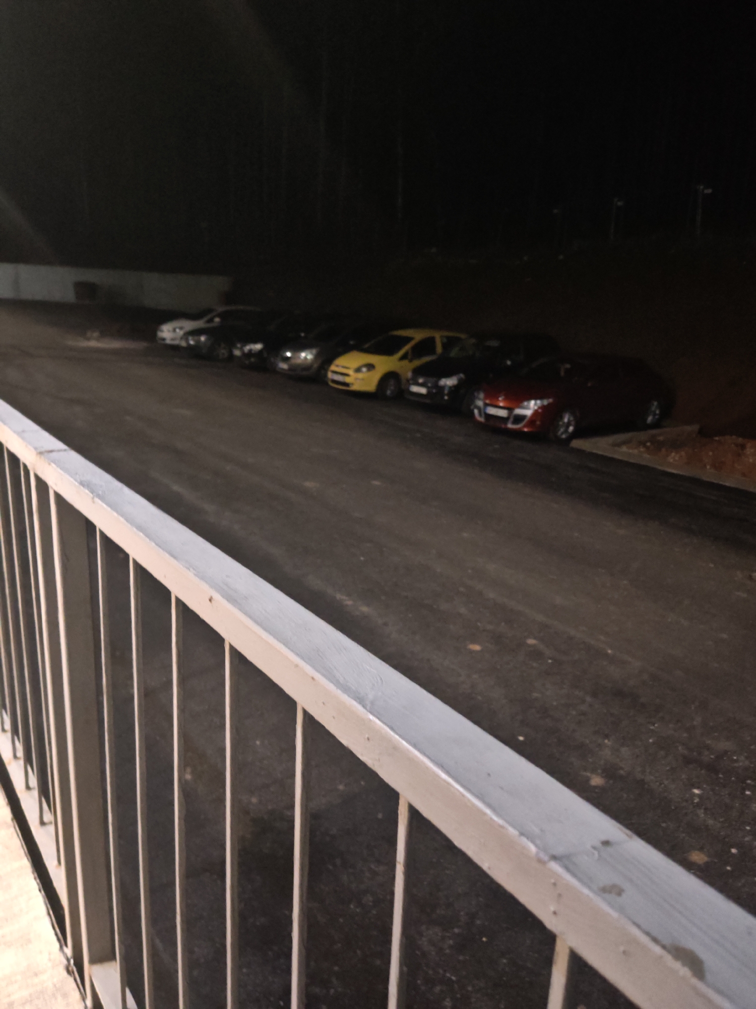 The image captures a night scene, likely a parking lot or area adjacent to a road. A series of parked cars are visible, lined up along the asphalt surface. The cars vary in color, including white, dark shades (possibly black or gray), yellow, and red. The area is illuminated by sporadic lighting, suggesting streetlights or nearby sources. A metal railing runs along the lower left edge of the frame, providing a foreground element. The scene is located in the city of Vratsa, Bulgaria, based on the available information. The environment is dark and slightly hazy.
