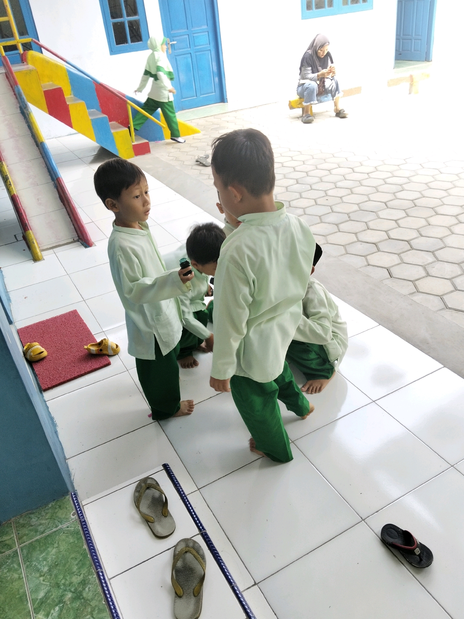 In Krajan Lor, Indonesia, a group of children in light green tops and dark green pants are gathered on a tiled outdoor patio. One child is holding a small dark object. Adjacent to them is a brightly colored slide with yellow, red, and blue sections, and a blue ramp. Further in the background, a woman wearing a headscarf and casual clothing sits on a yellow bench, looking at her phone. Another child in a similar outfit to the others is walking up the slide's ramp. The environment appears to be a school or play area with white walls and blue doors in the background. Scattered on the tiles are several pairs of flip-flops and sandals, including yellow sandals near a red mat. The ground is paved with light-colored hexagonal and square tiles.