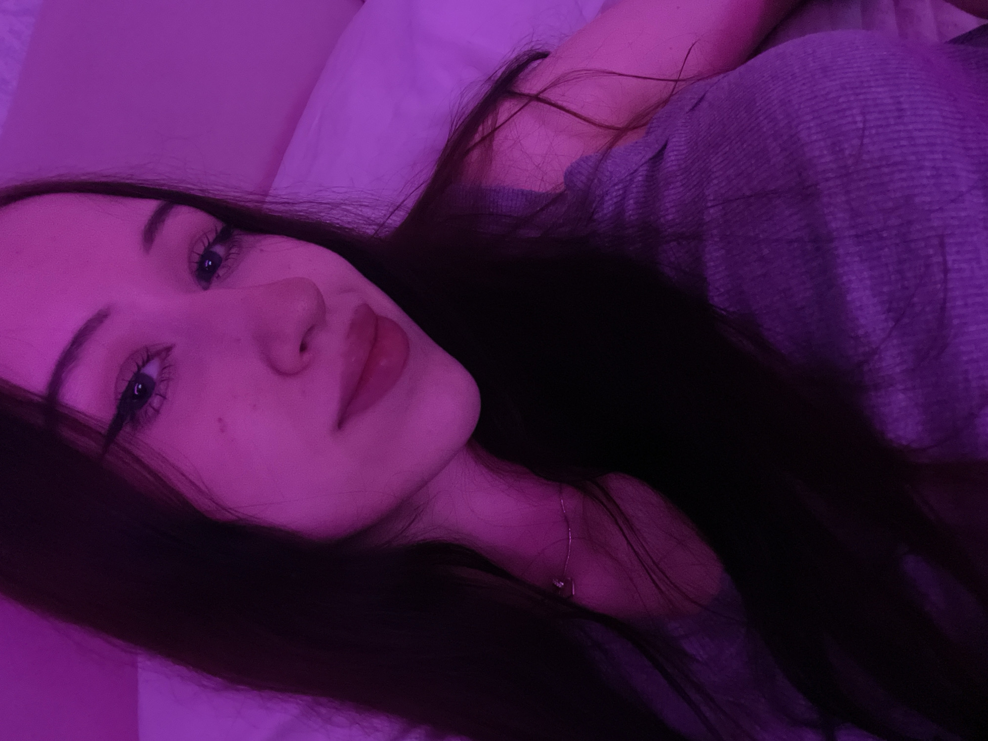 The image shows a close-up, waist-up shot of a young woman with long, dark hair, lying down in bed. She is wearing a grey ribbed tank top and a delicate silver necklace with a small pendant. The room is illuminated by pink-purple lighting, casting a neon glow over her skin and surroundings. She has noticeable eye makeup, including winged eyeliner, and her lips are painted a vibrant pink. Her expression is relaxed, with her head tilted slightly and a gentle smile on her face. The background consists of a white pillow and bedding, suggesting an indoor setting, likely a bedroom. There are no visible indications of Votkinsk, Russia, or any specific location within the city. The image appears to be a selfie taken in a private, dimly lit environment.