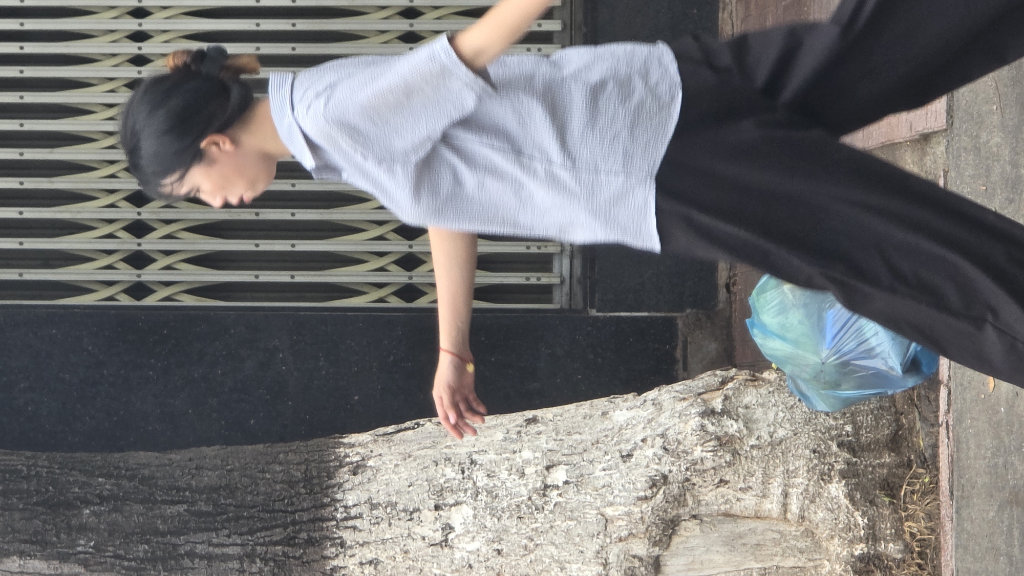 The image, rotated 90 degrees counter-clockwise, depicts a candid moment in an urban environment, likely in Bac Nha Trang Ward, Vietnam. The main subject is a young person, likely female, with dark hair tied back in a bun, who appears to be leaning or reaching downwards. She is dressed in a light-colored, possibly light blue or white striped, short-sleeved collared shirt and black trousers. A red string bracelet with a small gold-colored charm is visible on her right wrist. Her head is bowed, and her face is in profile, looking down towards a large tree trunk that dominates the lower half of the frame. Her right hand is extended, seemingly touching or hovering near the tree's textured bark, which features light gray and darker areas.
In the background, a metal rolling security shutter with a distinctive crisscross and horizontal line pattern is visible, suggesting a storefront or building facade. Below the shutter and behind the person is a dark, smooth surface, possibly concrete or tiled. To the right of the tree trunk and near the edge of what appears to be a sidewalk or ground cover, a light blue plastic bag is visible. The scene is illuminated by soft, diffused daylight, indicating it is daytime, possibly with overcast conditions. There is no visible text in the image.