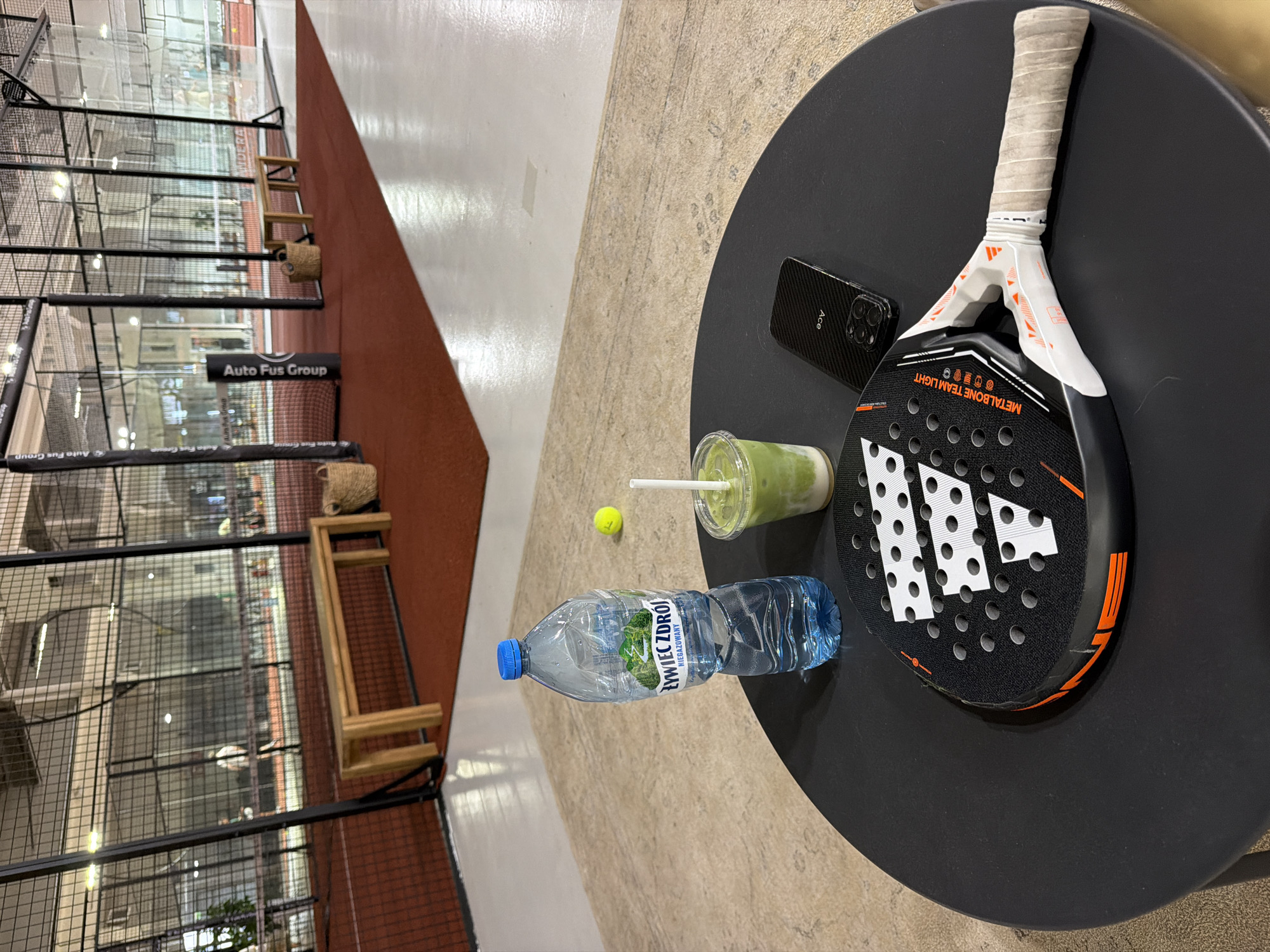 The image shows a table next to a padel court. On the table, there is a padel racket, a bottle of water, a smoothie, a yellow ball, and a phone. The padel court is visible in the background. The Auto Fus Group sign is visible on the wall of the court.