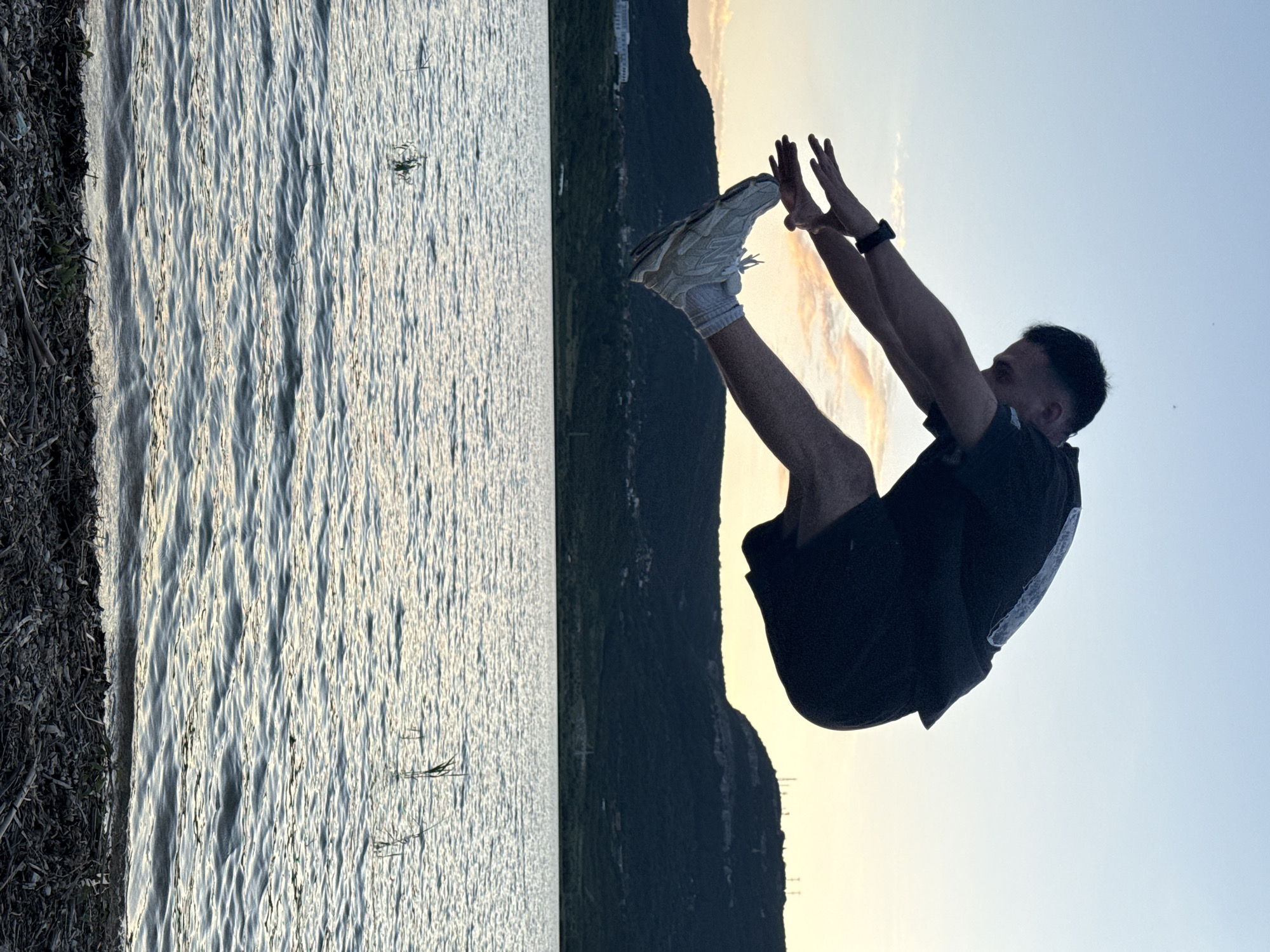 A man is captured mid-air, performing a jump over a body of water. He is wearing a dark t-shirt, dark shorts, white socks, and light-colored athletic shoes. His arms are extended forward, and his legs are bent. The scene appears to be set at dusk, with a hazy sky transitioning from light blue to pale yellow. In the background, a dark, forested hill with scattered structures and antennas rises above the water. The water's surface is rippled, reflecting the soft light of the sky. The foreground consists of a rocky shoreline with scattered twigs and vegetation. The image was taken in the city of Shumen, Bulgaria, near the Shumen Plateau Nature Park, which features a lake and hilly terrain.