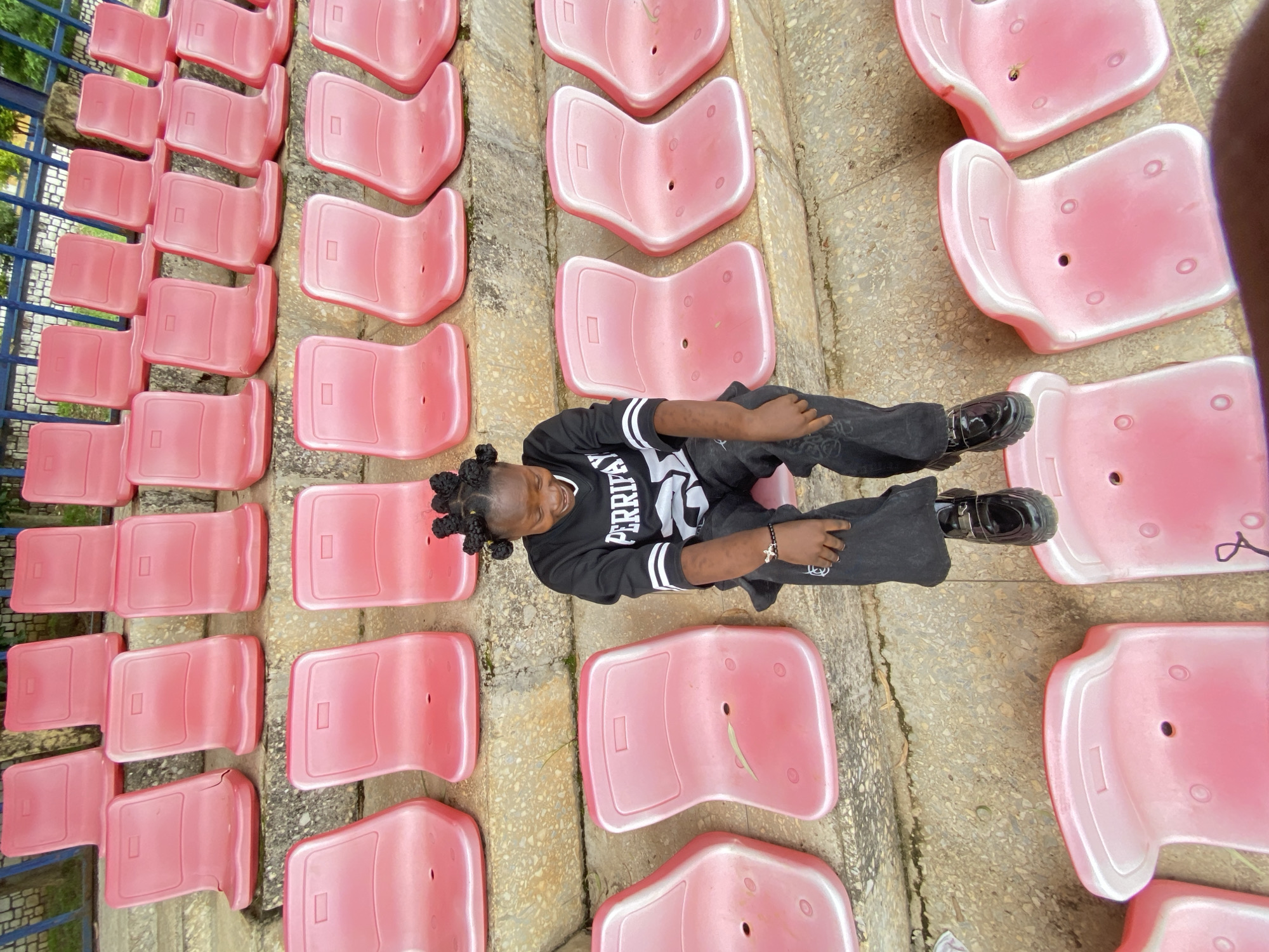 In Zawan, Nigeria, a young girl with braided hair and wearing a black sweatshirt with white lettering and black pants sits on a row of empty pink stadium seats. The seats are arranged in multiple tiers, and some are empty while others are occupied by the girl. The girl is looking down and smiling, with her left hand resting on her knee. The background shows a fence and some greenery. The scene appears to be outdoors, possibly at a sporting event or a public gathering. The lighting suggests it is daytime.