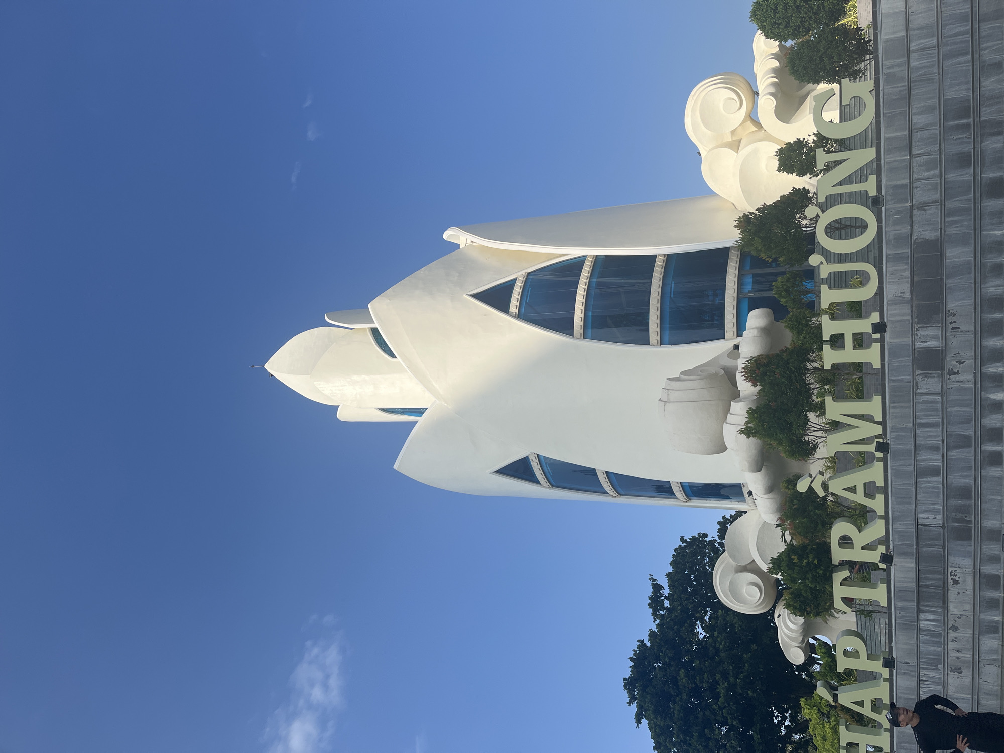 This is a low-angle, medium shot of the Phu Dong Thien Vuong tower in Nha Trang, Vietnam, on a clear, sunny day. The tower, a modern structure with a white, petal-like exterior and large, blue, triangular windows, dominates the frame. In the foreground, large, light green letters spell out "THÁP TRẦM HƯƠNG," indicating the name of the landmark. A lone individual, a man dressed in black, stands on a gray, tiered staircase to the left of the letters, facing towards the tower. Behind the letters and the man, lush green trees and decorative cloud-like sculptures surround the base of the tower. The sky is a vibrant, cloudless blue, with only a faint wisp of a cloud visible on the left side. The scene depicts a well-maintained public space, likely a tourist attraction, under bright daylight conditions.