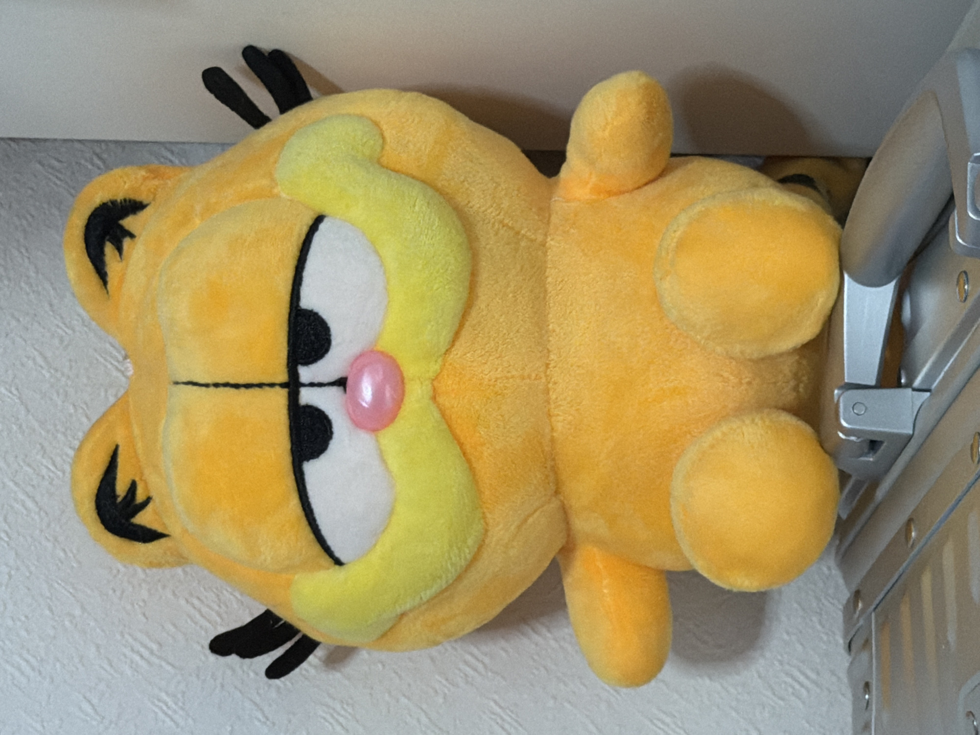 A plush toy of the character Garfield is positioned in the foreground of the image. The toy is predominantly orange-yellow, with black features on its ears and whiskers. It has large, closed eyes with white sclera and black pupils, and a pink, bulbous nose. The toy is seated on a metallic surface with visible rivets and indentations, which appears to be a part of a larger object. The background consists of a textured, light-colored wall. The scene is indoors, and there is no information available to determine its specific location within the city of Moscow, Russia.