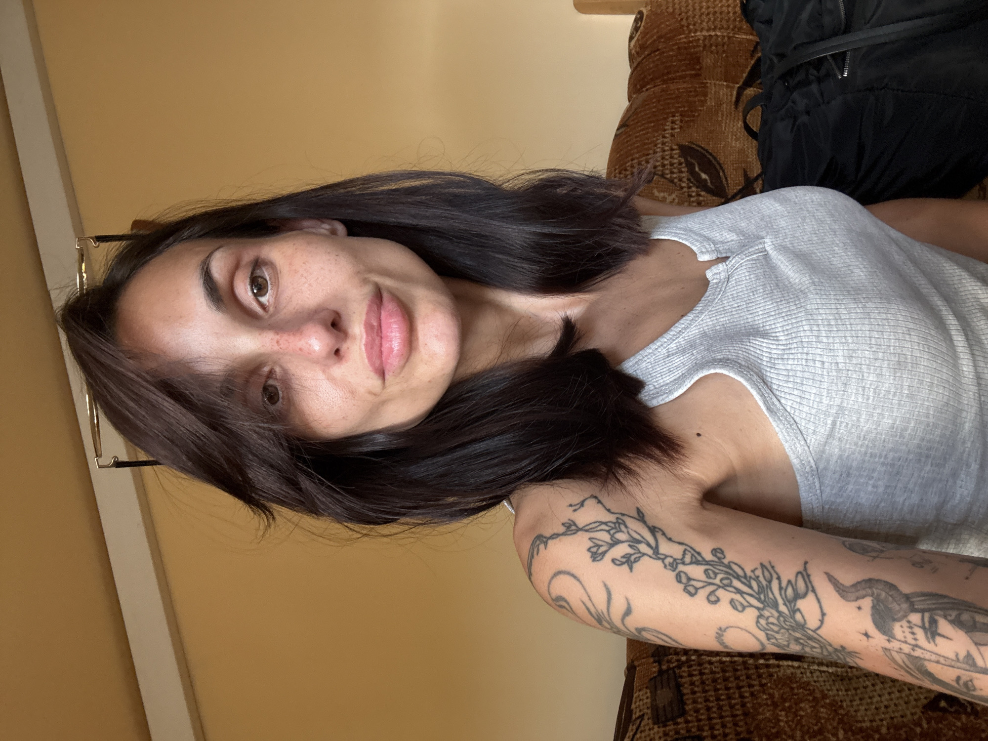 A medium, eye-level shot presents a woman with light skin, brown eyes, and dark brown hair, with a few strands falling across her face. She is wearing a light gray tank top. Visible on her arm is a detailed tattoo featuring floral and demonic elements. Behind her is a dark-colored bag or backpack resting on what appears to be a brown patterned sofa. The background also includes a light yellow wall. The image does not show any information indicating the location of Pleven, Bulgaria.