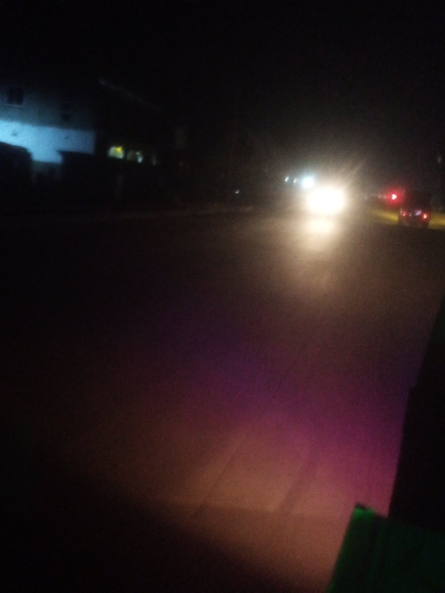 A nighttime street scene in Tundun Wada South, Nigeria, captured from a moving vehicle. The foreground is dark, with a blurry green object on the right side. The middle ground shows a road illuminated by headlights of an approaching car, casting a bright white light. The road also reflects purple and orange hues from ambient light sources. In the background, a building with illuminated windows is visible on the left, and taillights of another vehicle are present on the right. The overall environment suggests a suburban or residential area at night.