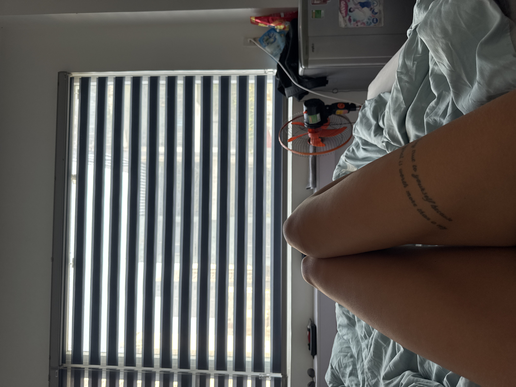 The image is taken indoors, presumably in a bedroom in Đà Nẵng, Vietnam. A person is lying in bed, with their legs crossed and visible in the foreground. The person has a circular tattoo on one of their legs. Behind the person is a bed covered with a light blue blanket. To the left of the bed is a window covered with striped blinds. To the right of the bed is a small refrigerator. A small fan is on the wall between the refrigerator and the window. Snacks can be seen on top of the fridge.